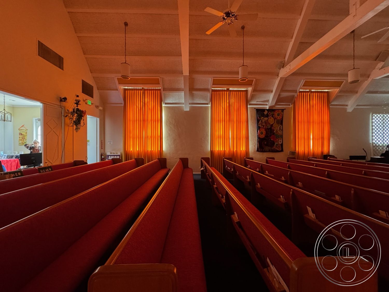 Spiritual Center - textured plaster walls, natural light from windows, bright ambient lighting, communal gathering area, exposed wood beam ceiling, architectural ceiling details, acoustic wall treatments, suspended ceiling fans, wooden pews, red upholstery seating, orange curtain panels, floral wall tapestry, church worship space