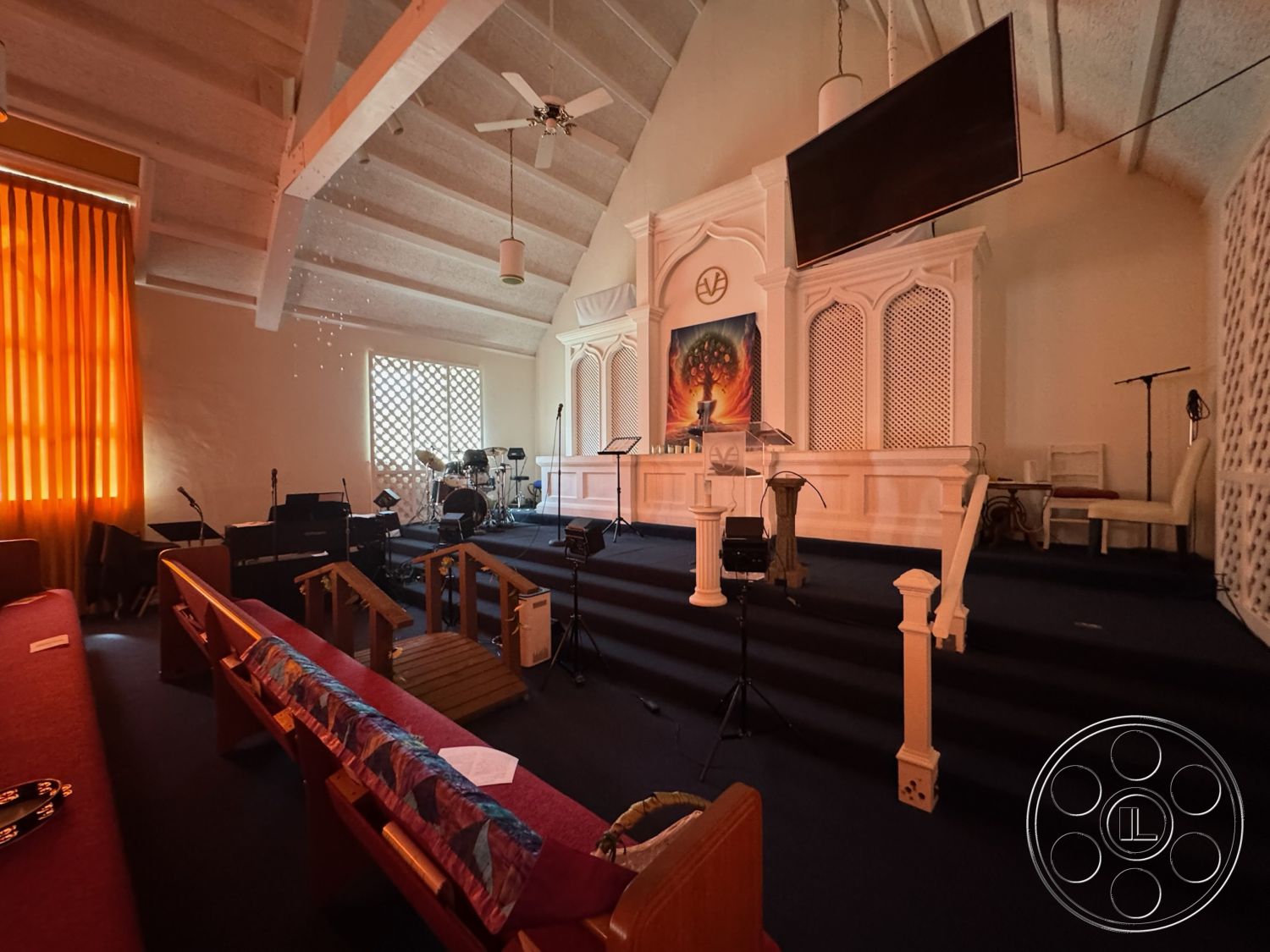 Spiritual Center - exposed wood ceiling beams interior, textured plaster walls interior, carpet flooring interior, vaulted ceiling interior, orange sheer curtain window treatment interior, stage area for performances interior, wooden pulpit interior, wooden benches seating interior, glass window with lattice design interior, modern sound equipment setup interior, artistic wall mural feature wall interior, acoustic paneling details interior, multicolored fabric accents on benches interior, spotlights for stage lighting interior, drum set band equipment area interior