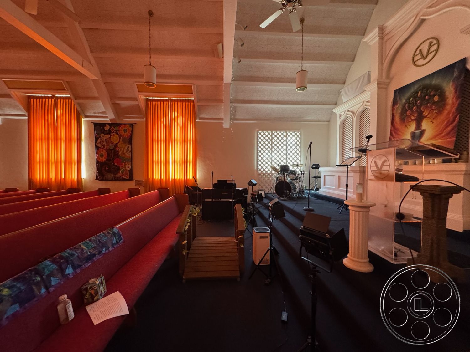 Spiritual Center - carpeted flooring, exposed wooden ceiling beams, wooden stage platform, acoustic drum set, interior worship space, orange patterned drapery, glass podium, colored artwork display, padded pew seating, abstract wall mural, architectural wood lattice, natural light filtering through windows, pulpit area with decorative columns, technical sound setup, multi-purpose gathering space