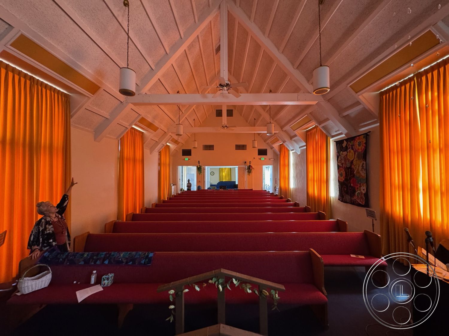 Spiritual Center - neutral wall color, ambient lighting fixtures, exposed wood ceiling beams, textured ceiling finish, wood paneling accent wall, wooden pews, cathedral ceiling, orange fabric curtains, rectangular sanctuary space, natural lighting from windows, entrance area with visible doorway