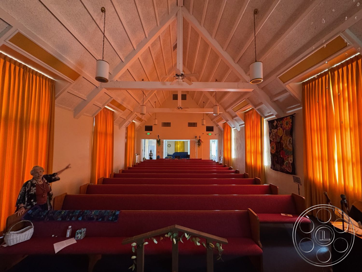 Spiritual Center - wood beam ceiling interior, high vaulted ceiling interior, red upholstered church pews interior, orange sheer curtains interior, white textured walls interior, hanging pendant lights interior, bright multi-colored wall tapestry interior, spacious worship area interior, natural daylight illumination interior, open congregational space interior