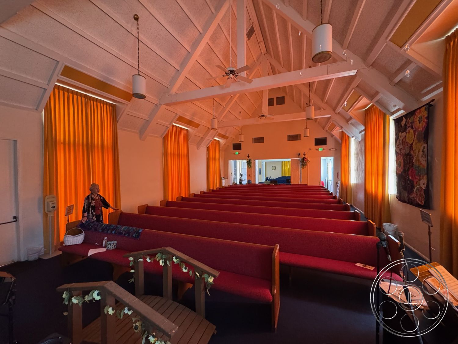 Spiritual Center - exposed wood beam ceiling interior, textured plaster walls interior, natural light from windows interior, high vaulted ceiling interior, orange curtain drapery interior, red upholstered pew seating interior, wooden stage platform interior, soft lighting fixtures interior, colorful textile wall hanging interior, acoustic ceiling tiles interior, open layout space interior, wooden entryway doors exterior
