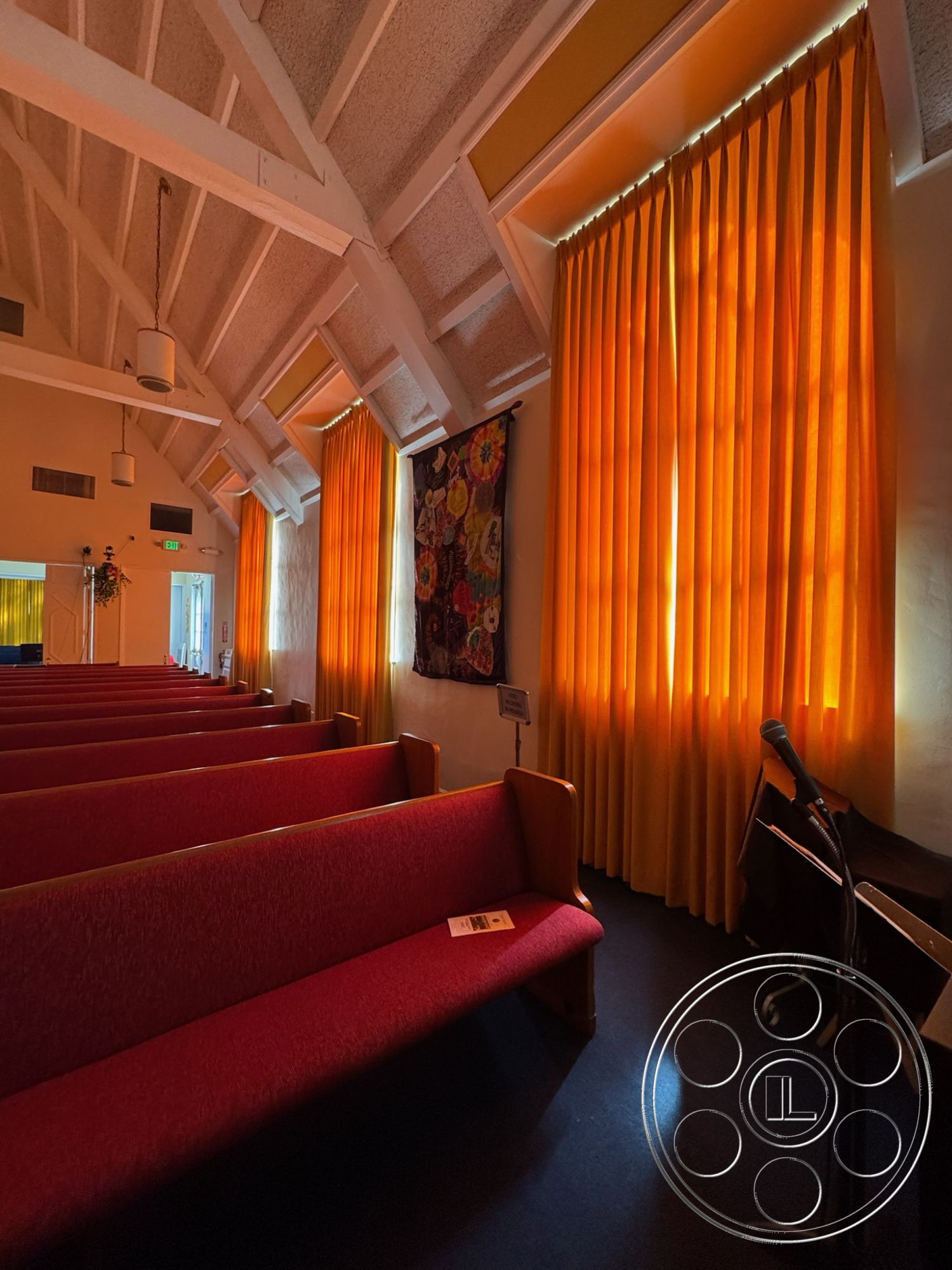 Spiritual Center - natural light from windows, exposed wood ceiling beams, textured plaster wall, indirect lighting fixtures, vaulted ceiling interior, red upholstered pews, orange velvet curtain, wooden pew construction, softwood church interior, acoustic paneling details, flooring with dark finish, decorative fabric wall hanging, liturgical space design