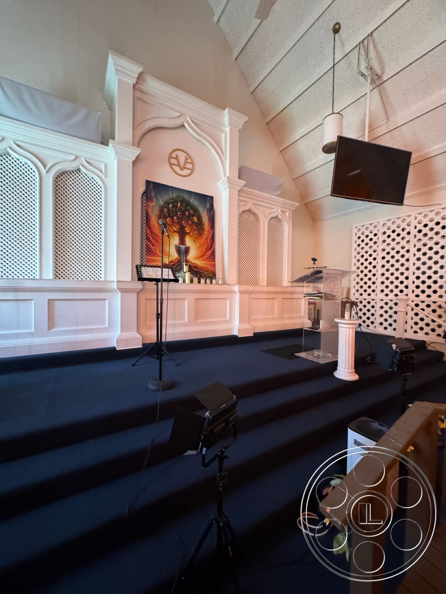 Spiritual Center - exposed wooden ceiling beams, interior sanctuary design, stained glass wall art, white wood architrave details, blue carpet flooring, painted wooden pulpit, decorative lattice side panels, golden decorative symbol, uplighting for stage area, moving spotlight fixtures, multicolored artwork installation, elevated platform steps, high vaulted ceiling with acoustic panels, minimalistic contemporary design, function for worship services