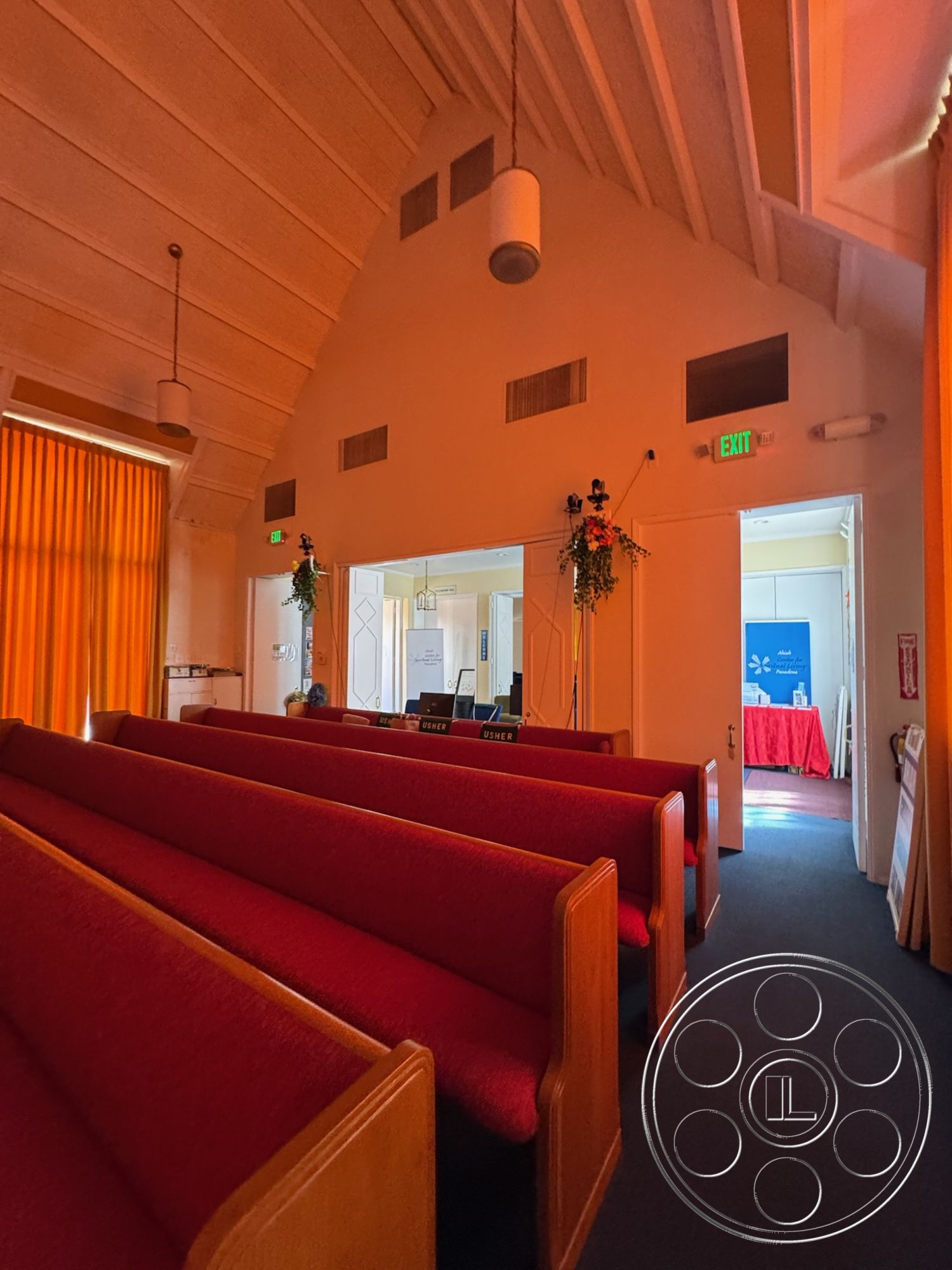 Spiritual Center - carpet flooring, vaulted ceiling, community gathering space, decorative wall molding, exposed wood beam ceiling, double door entrance, red upholstered church pews, wood paneling details, large windows with light-filtering curtains, traditional church architecture, brightly lit entryway, church sanctuary, ambient lighting from ceiling fixtures