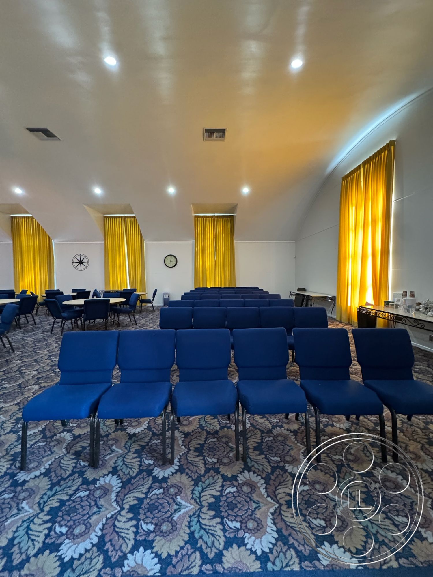 Spiritual Center - coffered ceiling, gathering space, blue upholstered chairs, arched ceiling, round tables, neutral painted walls, ambient recessed lighting, floral patterned carpet, metal chair legs, yellow drapery curtains, clock on wall, multipurpose room setup, community meeting area