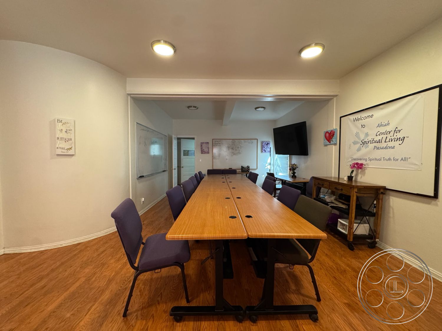 Spiritual Center - open floor plan, multifunctional space, decorative wall art, white painted walls, exposed ceiling lights, natural light windows, wooden conference table, purple upholstered chairs, light wood laminate flooring, dry erase board wall treatment, interior meeting room, circular room shape, modern meeting space, casual workspace ambiance