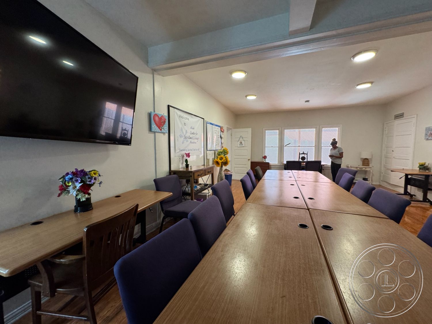 Spiritual Center - hardwood flooring, natural light from windows, exposed ceiling beams, multi-purpose room, textured plaster wall, white painted walls, interior conference room, formal meeting space, solid wood conference table, upholstered purple chairs, large wall-mounted flat-screen TV, decorative flower arrangements, double-door entry