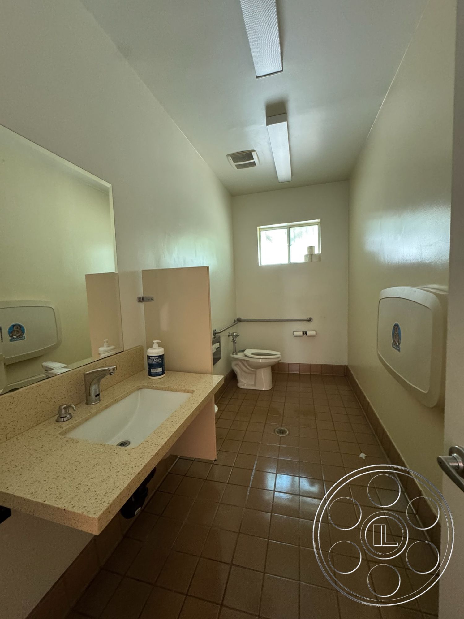 Spiritual Center - minimalist design, textured plaster walls, flush toilet, overhead fluorescent lighting, small window for natural light, white painted walls, tile bathroom flooring, pebble stone sink countertop, stainless steel faucet, rectangular sink, functional commercial bathroom layout, grab bars for accessibility, square mirror above sink, beige tiled walls, clean and modern aesthetics