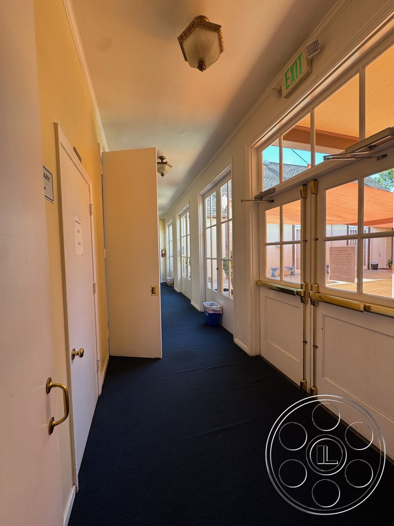 Spiritual Center - exit sign, soft natural lighting, open doorway, interior corridor, black carpet flooring, paneled doors, gold handle fixtures, walls painted light yellow, glass windows with white frames, traditional ceiling light fixture, white trim details, recycle bin, architectural transition space