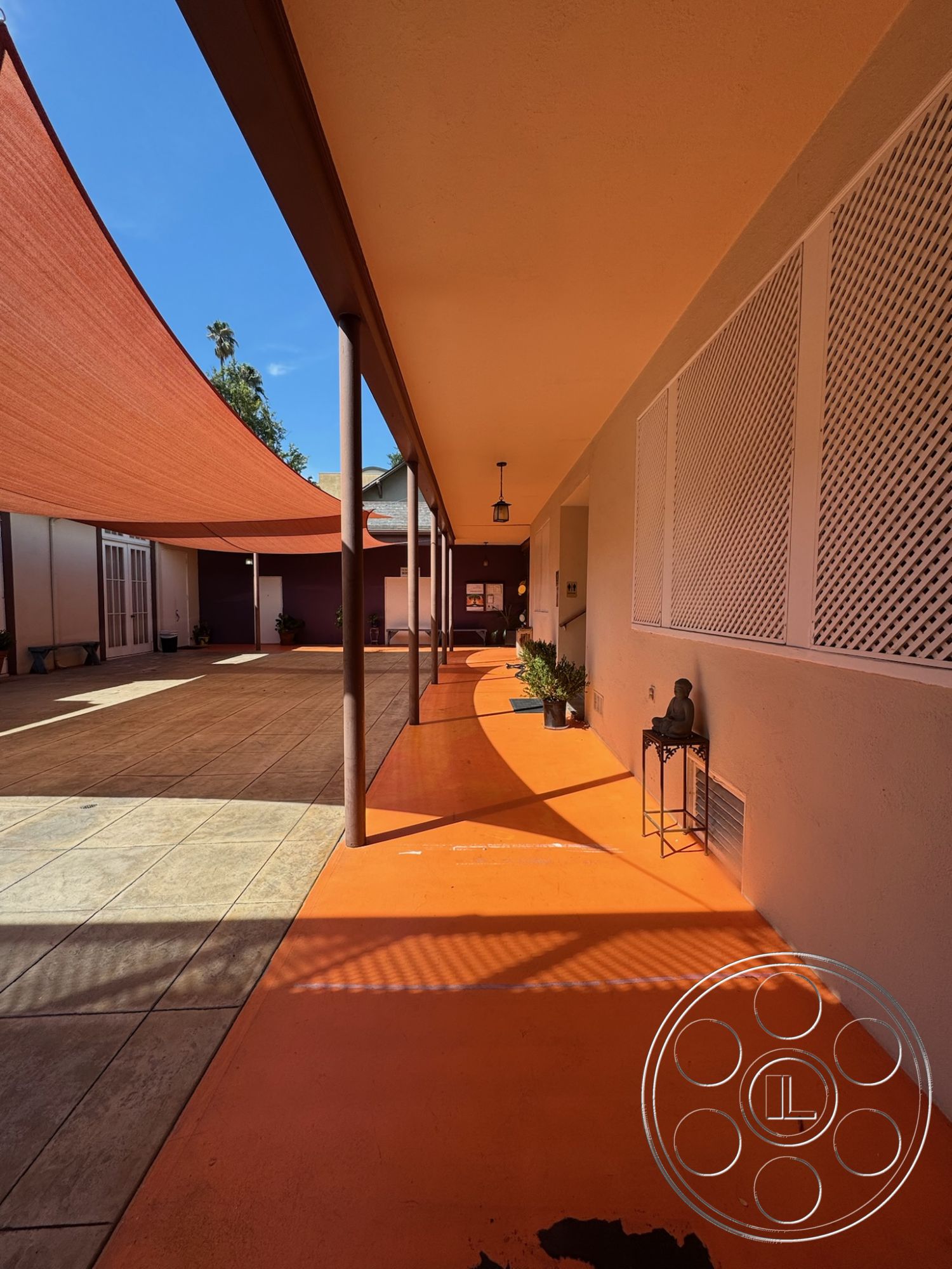 Spiritual Center - exterior textured plaster walls, exterior potted plants, exterior landscaping with trees, exterior orange concrete walkway, exterior shaded patio with sail shades, exterior stone tile courtyard flooring, exterior vertical slatted wood privacy screen, exterior decorative light fixture, exterior sunlit corridor