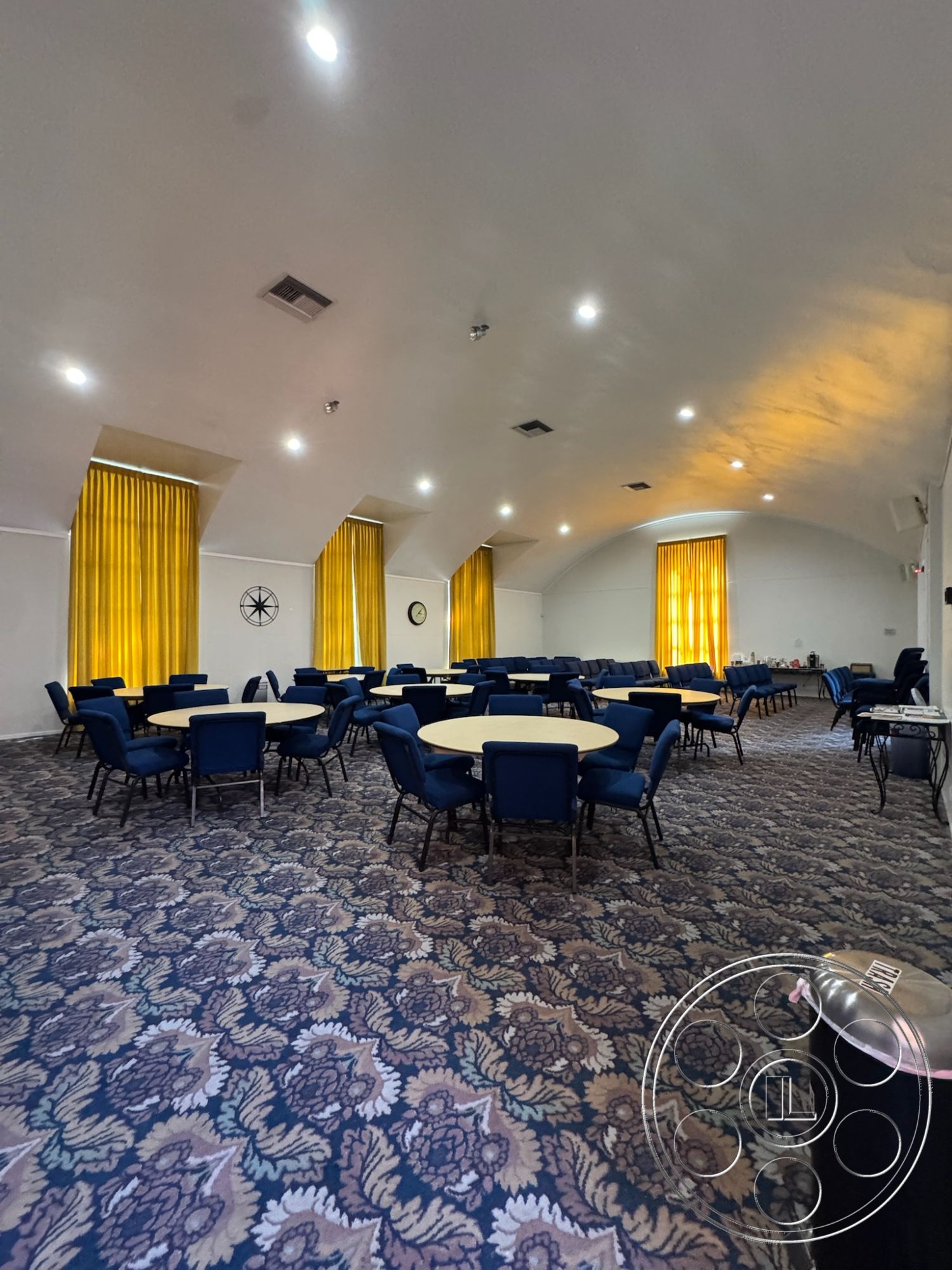 Spiritual Center - decorative wall clock, neutral painted walls, high ceilings with recessed lighting, acoustic ceiling treatment, interior event space, textured carpet flooring with floral pattern, round dining tables with blue fabric chairs, yellow drapery providing natural light control, clock wall accent, accessory table along the wall, open layout for flexible use, multi-purpose function room