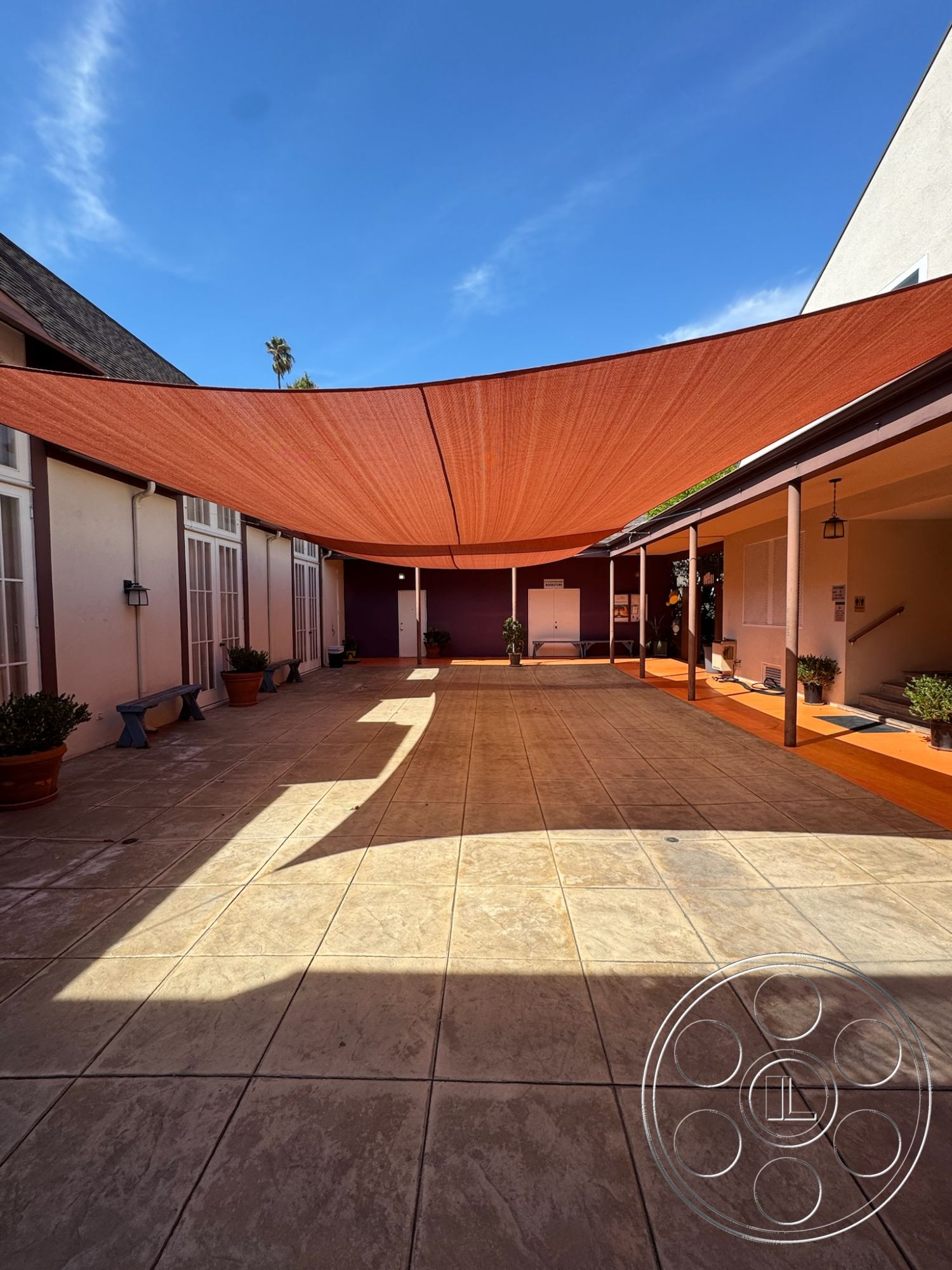 Spiritual Center - exterior, ambient natural lighting, modern outdoor area, bright blue sky, minimalist architectural design, potted plants accents, concrete planter boxes, rectangular courtyard layout, textured concrete patio flooring, orange shade sail, surrounding stucco walls, white wood-framed windows, shaded outdoor space