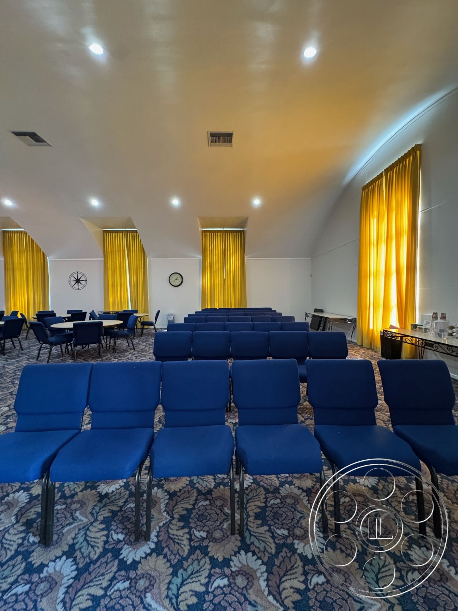 Spiritual Center - vaulted ceiling, blue upholstered chairs, decorative wall clock, open space for gatherings, recessed ceiling lights, neutral painted walls, interior multi-purpose room, acoustic ceiling design, metal chair legs, beige patterned carpet flooring, yellow sheer curtain drapes, round dining tables with chairs, wall-mounted service counter, decorative corner table