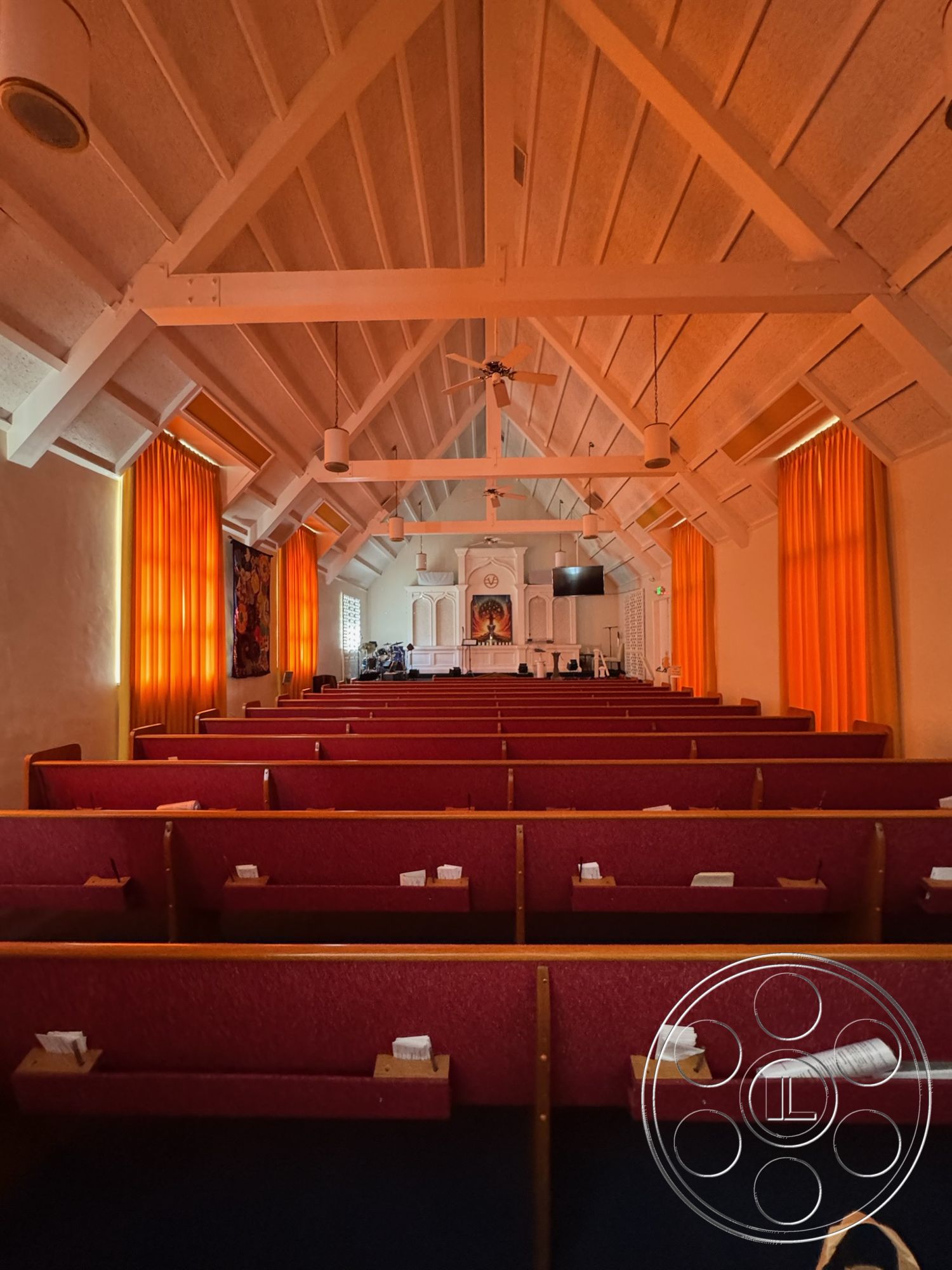 Spiritual Center - wood beam ceiling interior, gathering space interior, painted drywall walls interior, orange fabric curtain interior, padded red church pews interior, carpeted floor interior, floral wall artwork interior, traditional altar design interior, suspended ceiling fans interior, natural light through windows interior, wooden trim details interior, raised platform for altar interior, rectangular sanctuary layout interior, acoustic wall panels interior