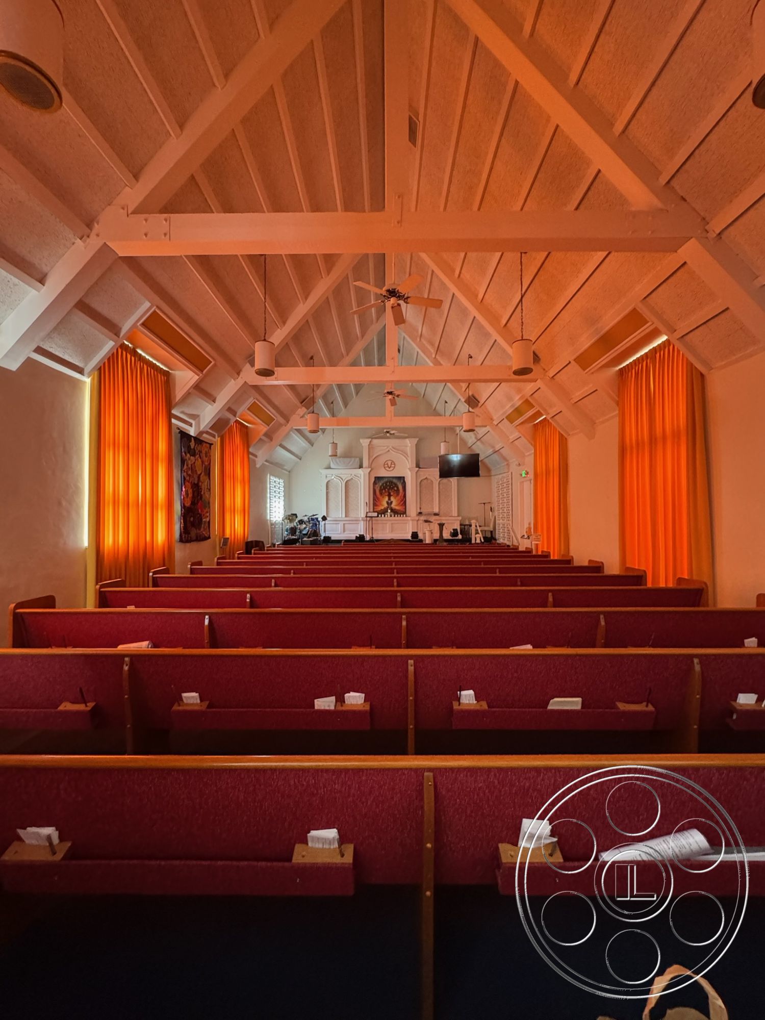 Spiritual Center - floor-to-ceiling windows, vaulted ceiling, multi-purpose space, neutral color scheme, ambient lighting fixtures, acoustic ceiling tiles, wood beam ceiling, open room layout, textured plaster wall treatments, historic architectural style, interior church space, orange sheer curtains, exposed wooden trusses, red fabric pew seating, raised pulpit area, white-painted wooden walls, congregational seating arrangement