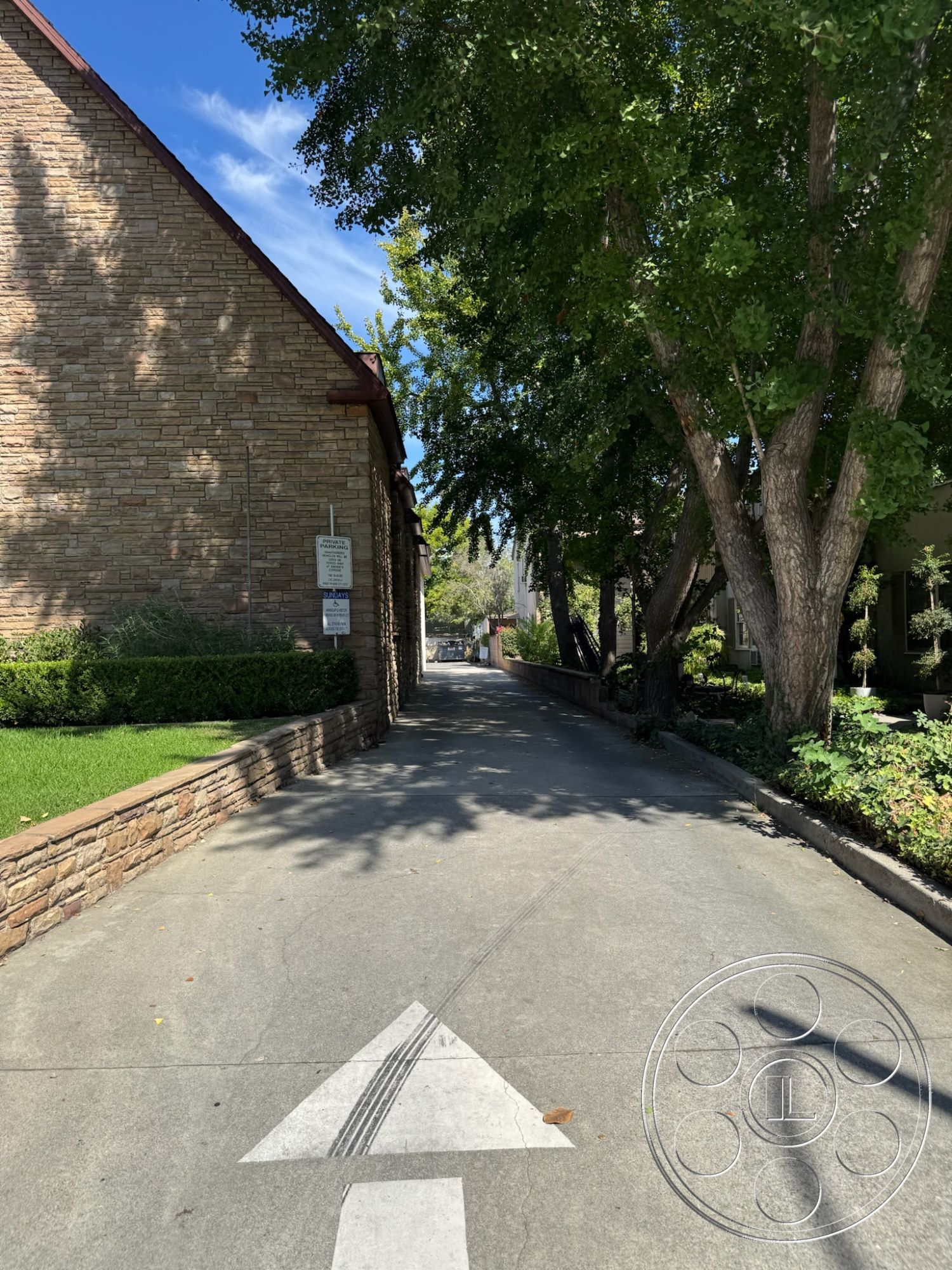 Spiritual Center - urban residential setting, exterior concrete driveway, hardscape pathway, asphalt parking area, exterior stone wall, landscaped garden edges, concrete pathway surface, textured stone border, street-facing tree canopies, well-maintained green lawns, sunlit outdoor environment, exterior architectural details, traditional stone construction, surrounding trees and foliage, clear blue sky backdrop, narrow passageway between buildings