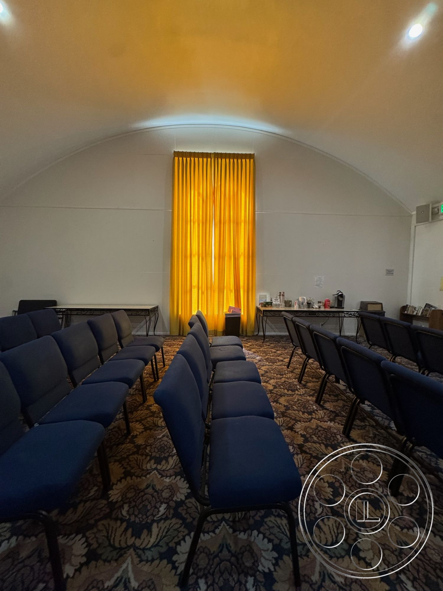 Spiritual Center - interior space, textured plaster walls, arched ceiling, patterned carpet flooring, yellow curtain treatment, navy blue seating, rectangle-shaped seating arrangement, community meeting room, natural lighting from window, refreshment area with tables