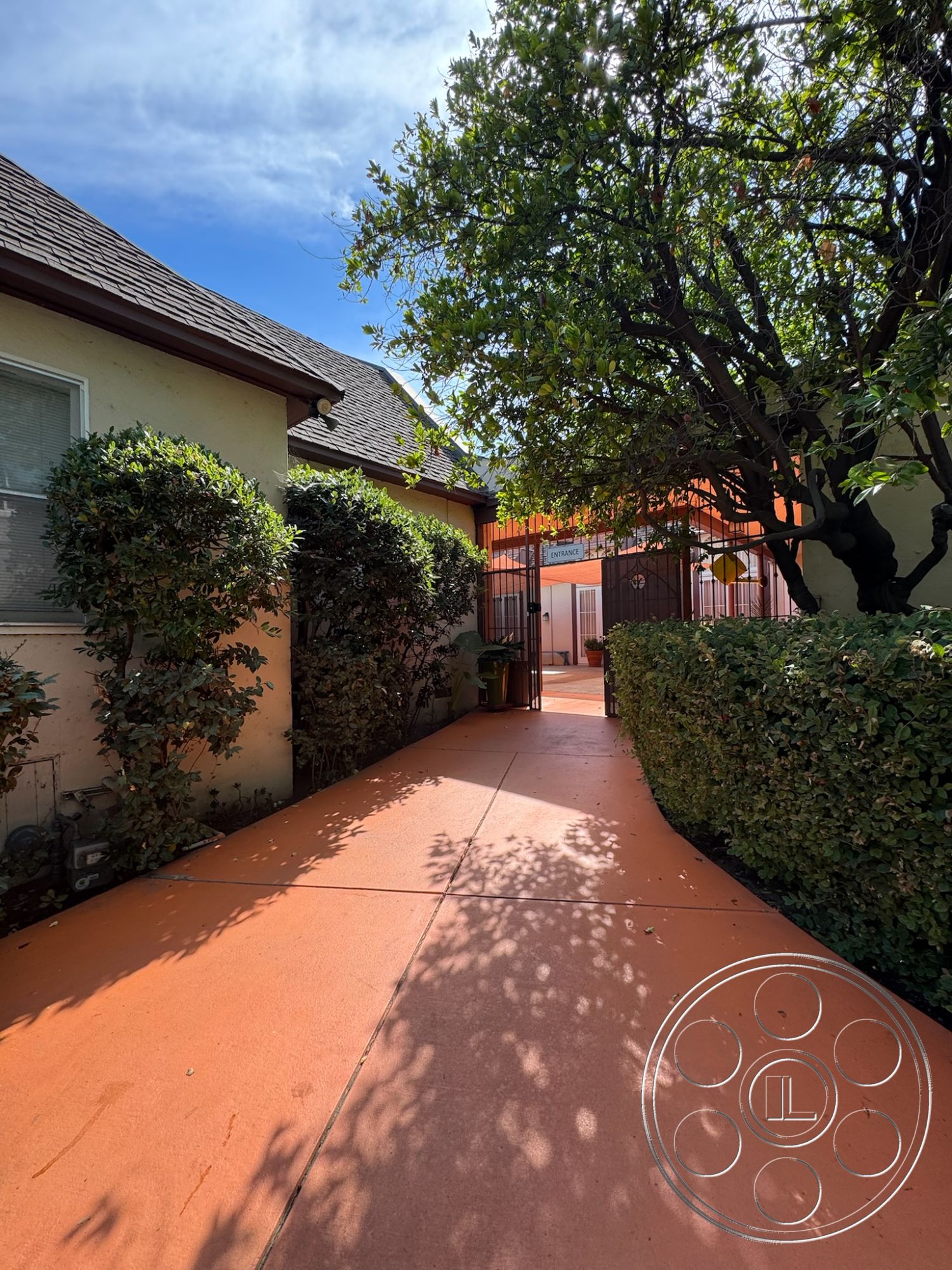 Spiritual Center - asphalt shingle roof, tree canopy, outdoor ambiance, sunlit pathway, landscaped hedges, urban garden setting, sun-drenched outdoor space, metal gate entrance, exterior orange concrete walkway, single-story residential home, framed entrance, surrounding shrubbery, shadow patterns on pavement