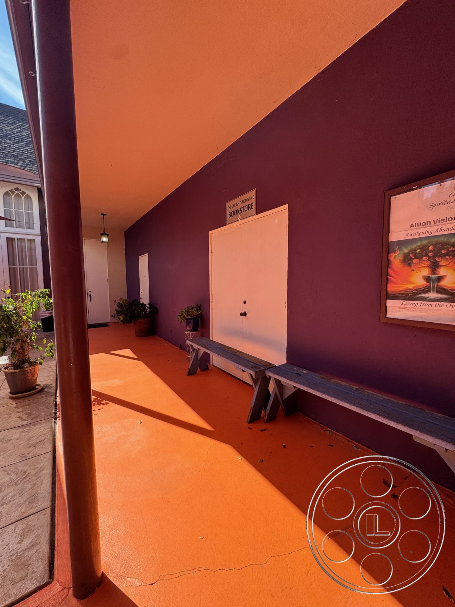 Spiritual Center - exterior wall treatments: purple painted wall, exterior flooring: textured orange concrete floor, exterior structural elements: round wooden support column, exterior benches: wooden plank benches, interior door features: solid white door, interior signage: bookstore sign, planting details: potted plants, setting: courtyard style, lighting: hanging pendant light fixture