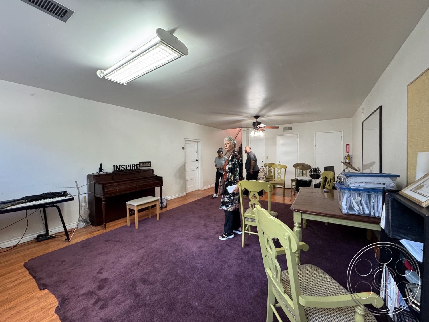 Spiritual Center - exposed wood ceiling beams interior, white painted walls interior, laminate wood flooring interior, purple carpet flooring interior, piano room with acoustic piano, brass ceiling fan interior, traditional wooden chairs interior, small wooden table interior, acoustic drum set interior, music studio space interior, inspire sign decor interior, home office workspace interior, multi-function room interior