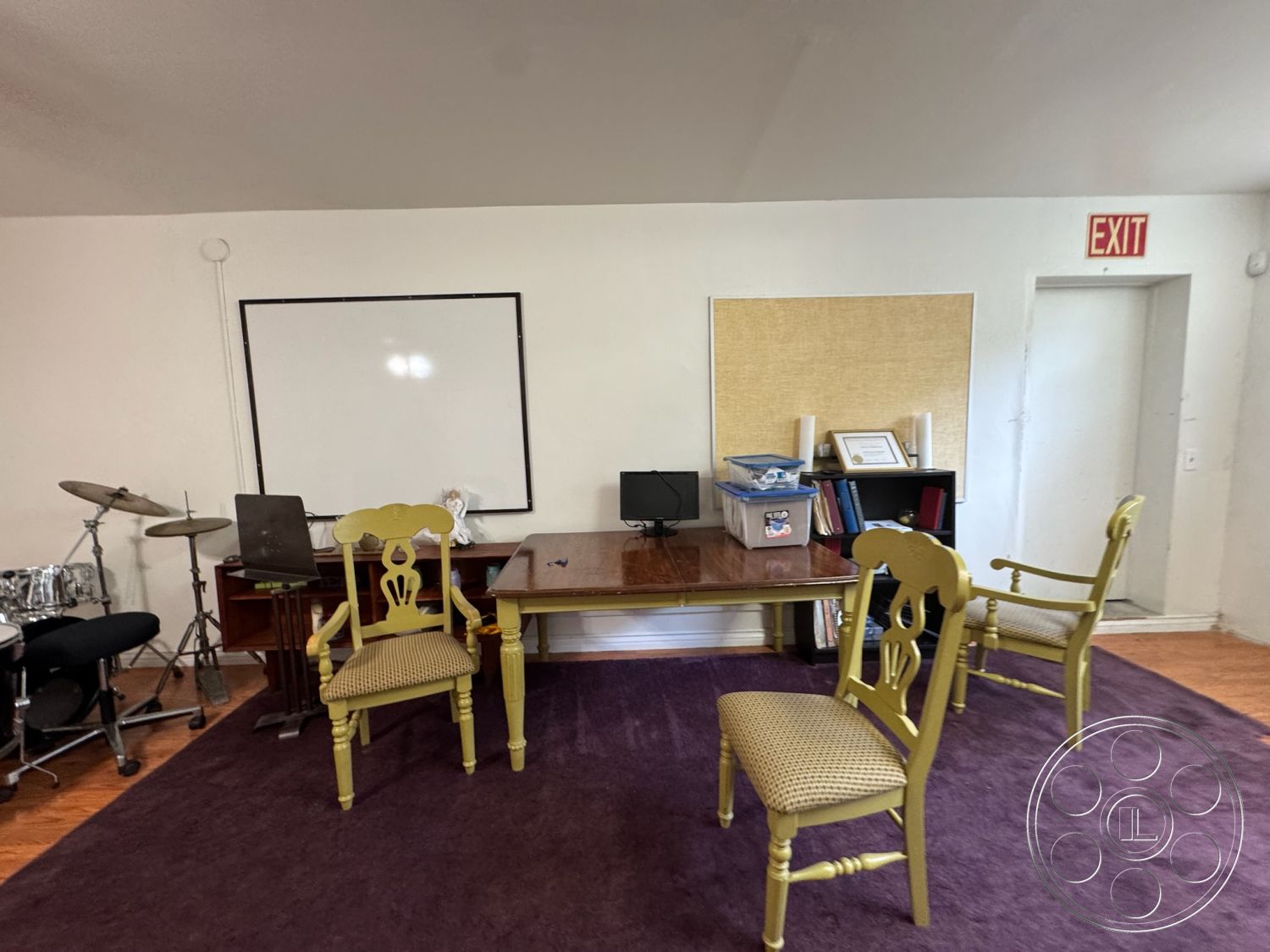 Spiritual Center - hardwood flooring, open floor plan, multifunctional space, wooden table, whiteboard wall, acoustic drum set, purple area rug, yellow painted chairs, acrylic wall art, shelving unit with books, natural light from door, exit sign above door, music practice area, relaxed meeting room, descriptive wall decor