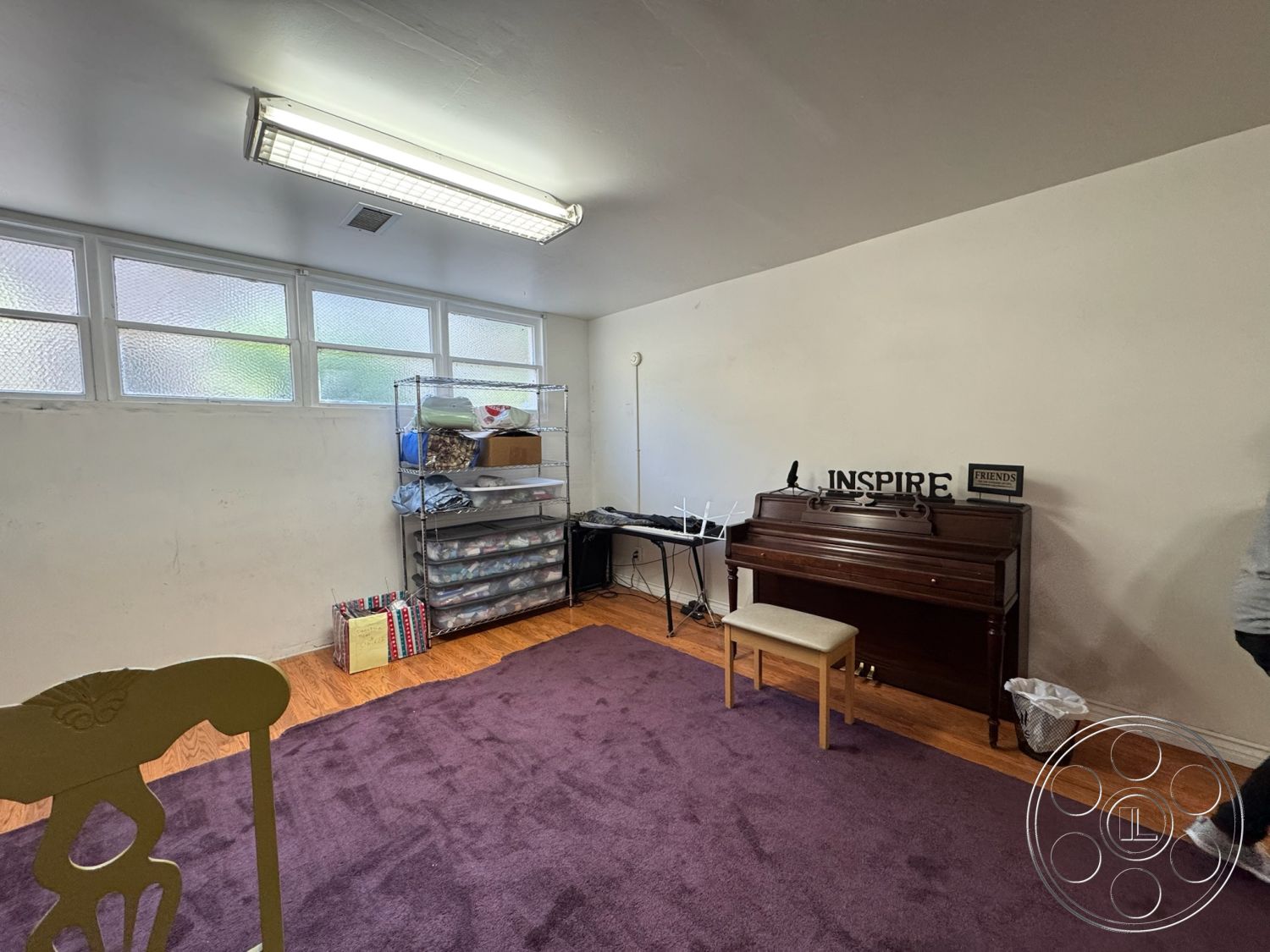 Spiritual Center - minimalist decor, open space design, workspace environment, glass block windows, light gray painted walls, purple area rug, laminate hardwood flooring, exposed fluorescent ceiling lights, wooden piano, white decorative accent wall, storage shelving unit, simple folding chair, compact seating area, utility room function