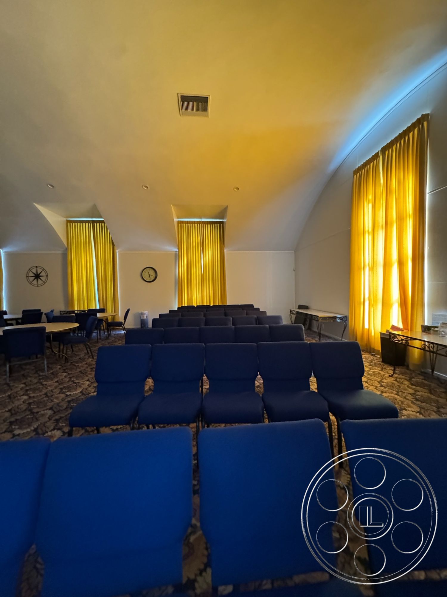 Spiritual Center - interior space, neutral wall color, high vaulted ceiling, large windows with natural light, functional meeting area, wall-mounted clock, yellow sheer curtains, blue upholstered seating, carpet flooring with floral pattern, modern table design, round tables for gatherings, ambient lighting with soft glow, open layout for versatile use, acoustic-friendly design