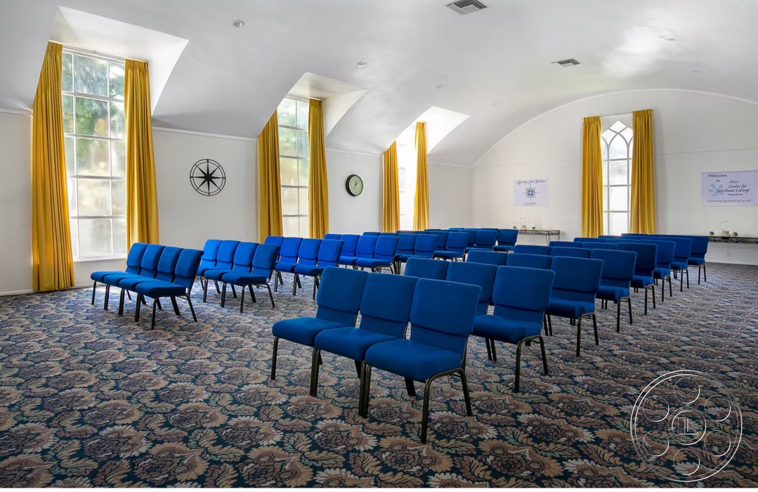 Spiritual Center - open layout, soft lighting, carpet flooring, neutral wall color, high vaulted ceiling, blue upholstered chairs, natural light from windows, large arched windows, decorative wall clock, floral patterned carpet, yellow drapery, minimalist wall decor, multipurpose gathering space, equipped with seating arrangements