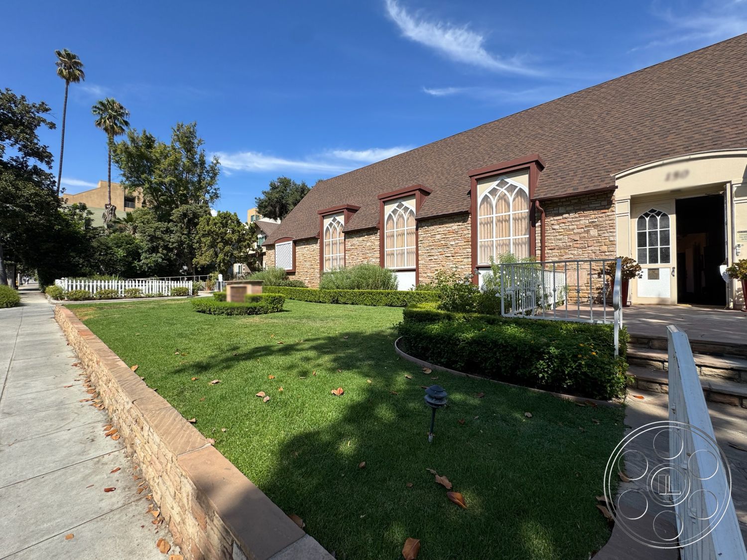 Spiritual Center - front porch, suburban setting, brick accent wall, concrete walkway, commercial property, palm trees in background, landscaped garden area, modern outdoor lighting fixtures, historic architectural style, gabled roof with shingles, exterior stone facade, arched window details, wooden handrail on steps, lawn area with ornamental shrubs, curved landscaping beds, easy access from sidewalk