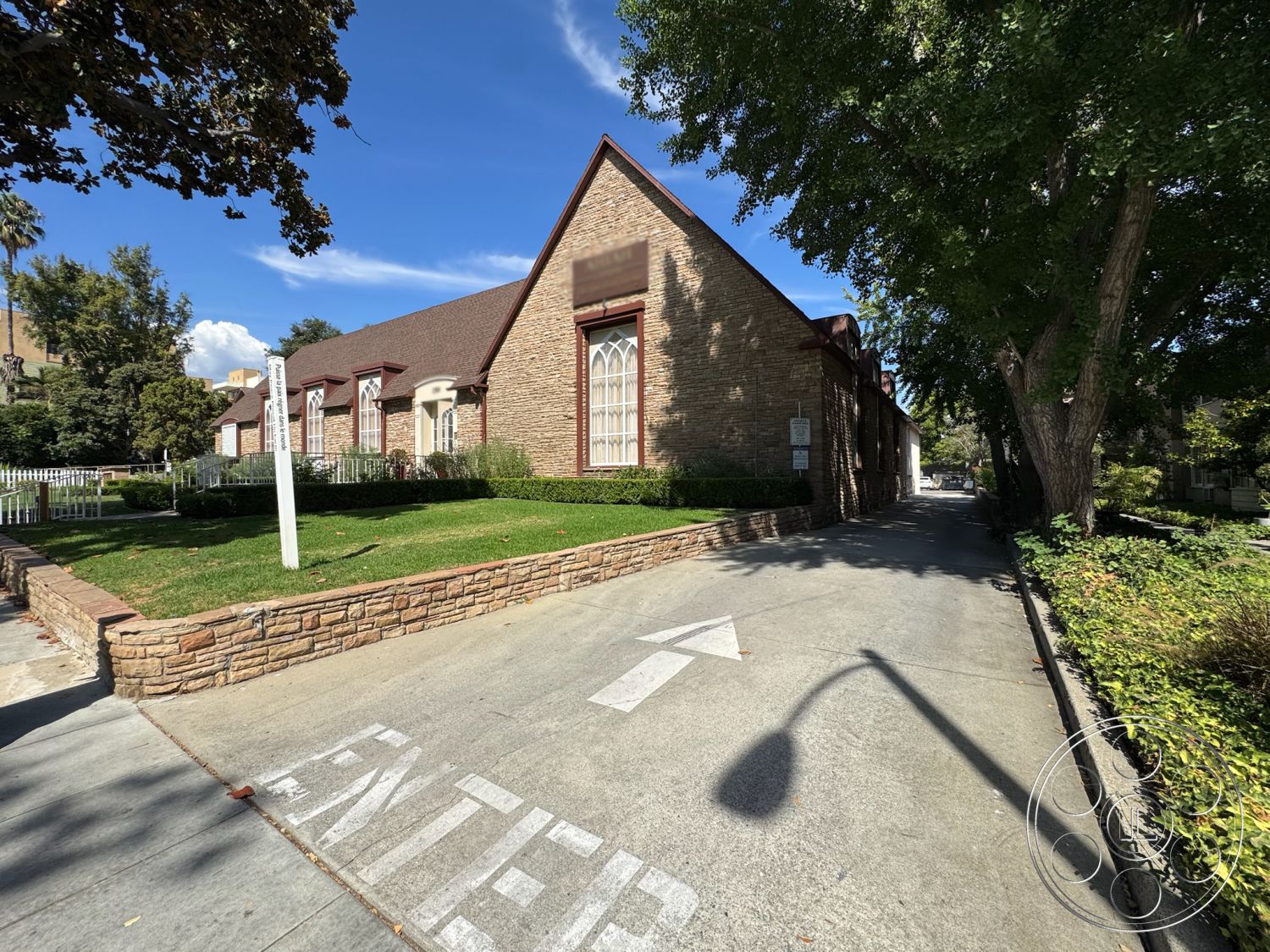 Spiritual Center - suburban setting, traditional architectural style, gabled roof shape, two-story building design, exterior stone brick walls, concrete driveway with directional markings, landscaped grass area with hedges, arched windows with decorative frames, low stone wall bordering the driveway, mature trees in surrounding landscape, asphalt shingle roofing, paved pathway leading to the entrance, decorative fence surrounding the property