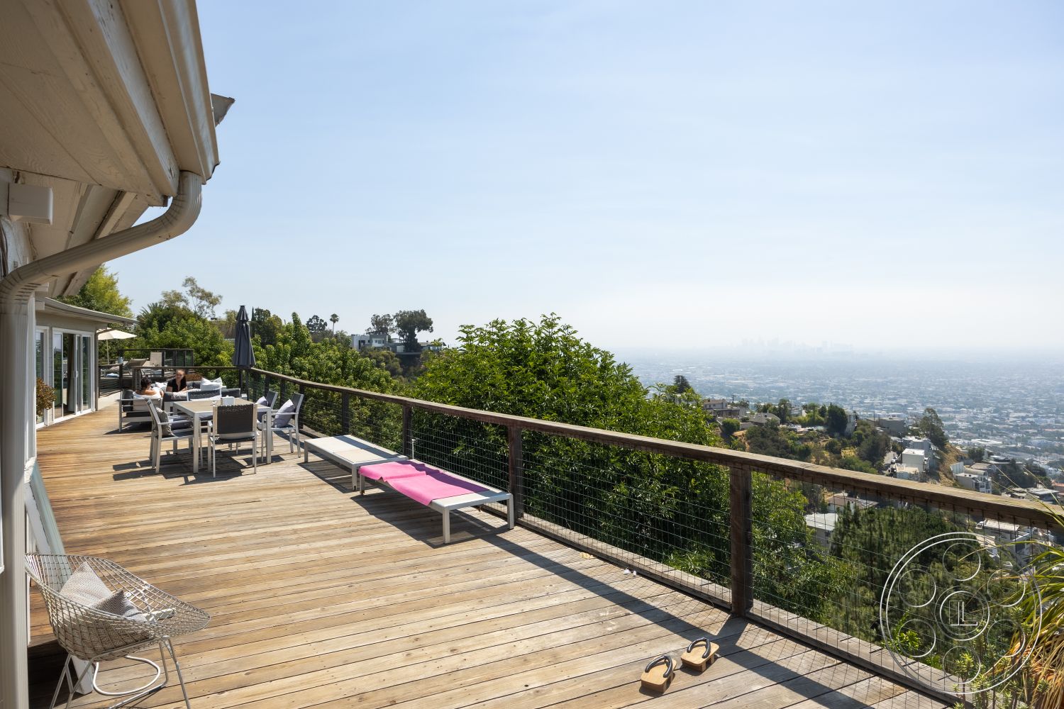 Retro 294 - natural wood finishes, comfortable seating arrangement, modern patio furniture, mid-century modern architectural style, minimalist decor elements, wood deck flooring, panoramic city skyline view, lush greenery surrounding the property, urban hillside setting, outdoor umbrella for shade, exterior railing with wire mesh, open outdoor dining area, sun lounger beside the deck, landscaped hillside garden, sloped lot with vegetation, covered outdoor dining space, iron support beams for the deck, deck space for entertaining, clear blue sky overhead