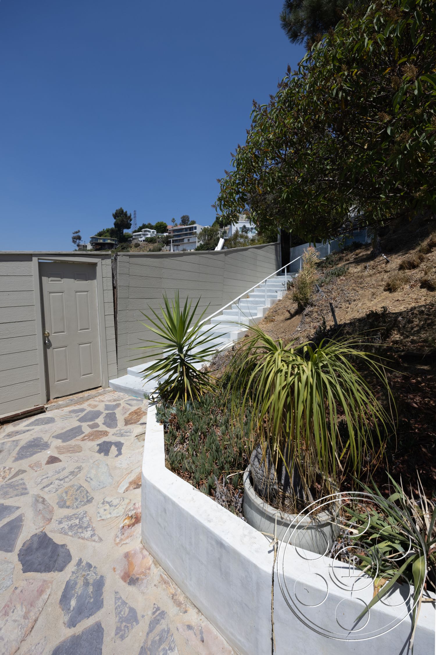 Retro 294 - landscaped garden area, minimalist architectural style, natural sunlight exposure, sloped hillside setting, outdoor stairway, exterior stone pathway flooring, irregular stone pavers, concrete flower bed border, light-colored concrete walls, succulent plants in planters, exterior wood door, urban hillside residence, desert landscape environment