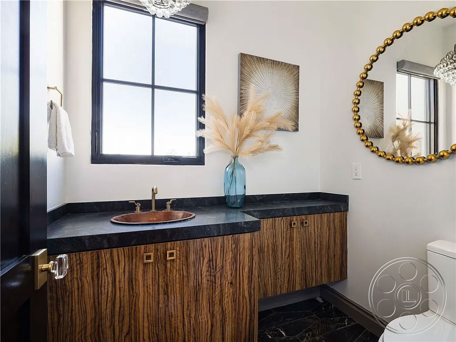 Spanish 167 - black granite countertop, interior bathroom design, neutral painted walls, contemporary bathroom style, wood veneer bathroom cabinetry, copper vessel sink, hexagonal black tile flooring, large round mirror with gold accents, modern minimalist lighting fixture, tall window with black frame, decorative pampas grass arrangement, gold hardware details
