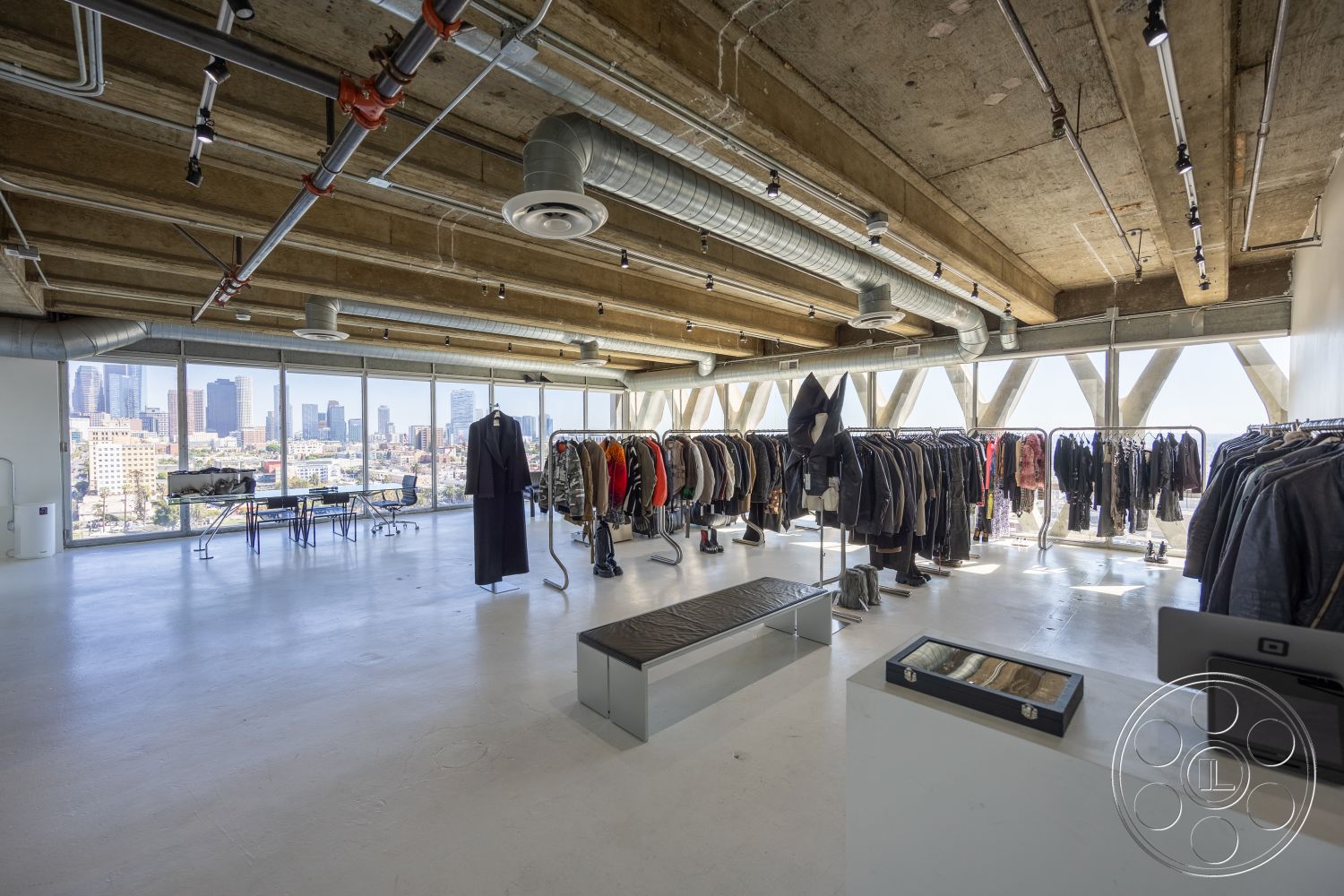 Loft 159 - neutral color palette, open floor plan, modern furniture, polished concrete flooring, functional workspace, industrial lighting fixtures, exposed ductwork, urban skyline view, bright and airy atmosphere, textured wall finishes, steel frame structure, floor-to-ceiling glass windows, minimalist interior, angular architectural shapes, contemporary retail space, metal clothing racks, concrete ceiling panels, city loft setting, cozy fitting area, large display windows