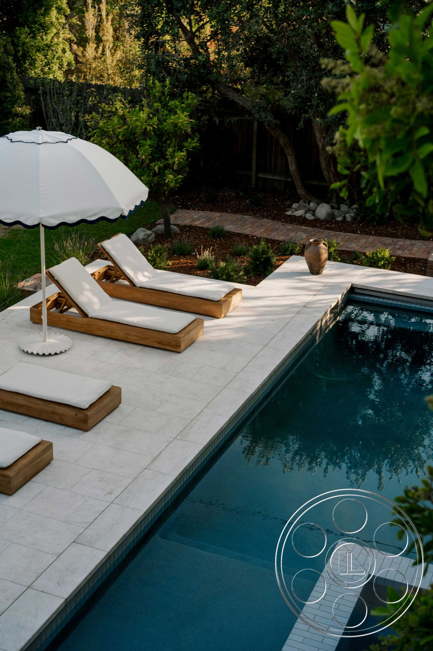 Americana 429 - rectangular swimming pool, brick pathway, sunbathing area, in-ground swimming pool, wooden lounge chairs, landscaped garden area, lush greenery surrounding the pool, natural stone planter, modern outdoor living space, exterior concrete pool decking, marble patio flooring, white umbrella with navy trim, exposed wooden elements, clear water reflection, outdoor relaxation zone