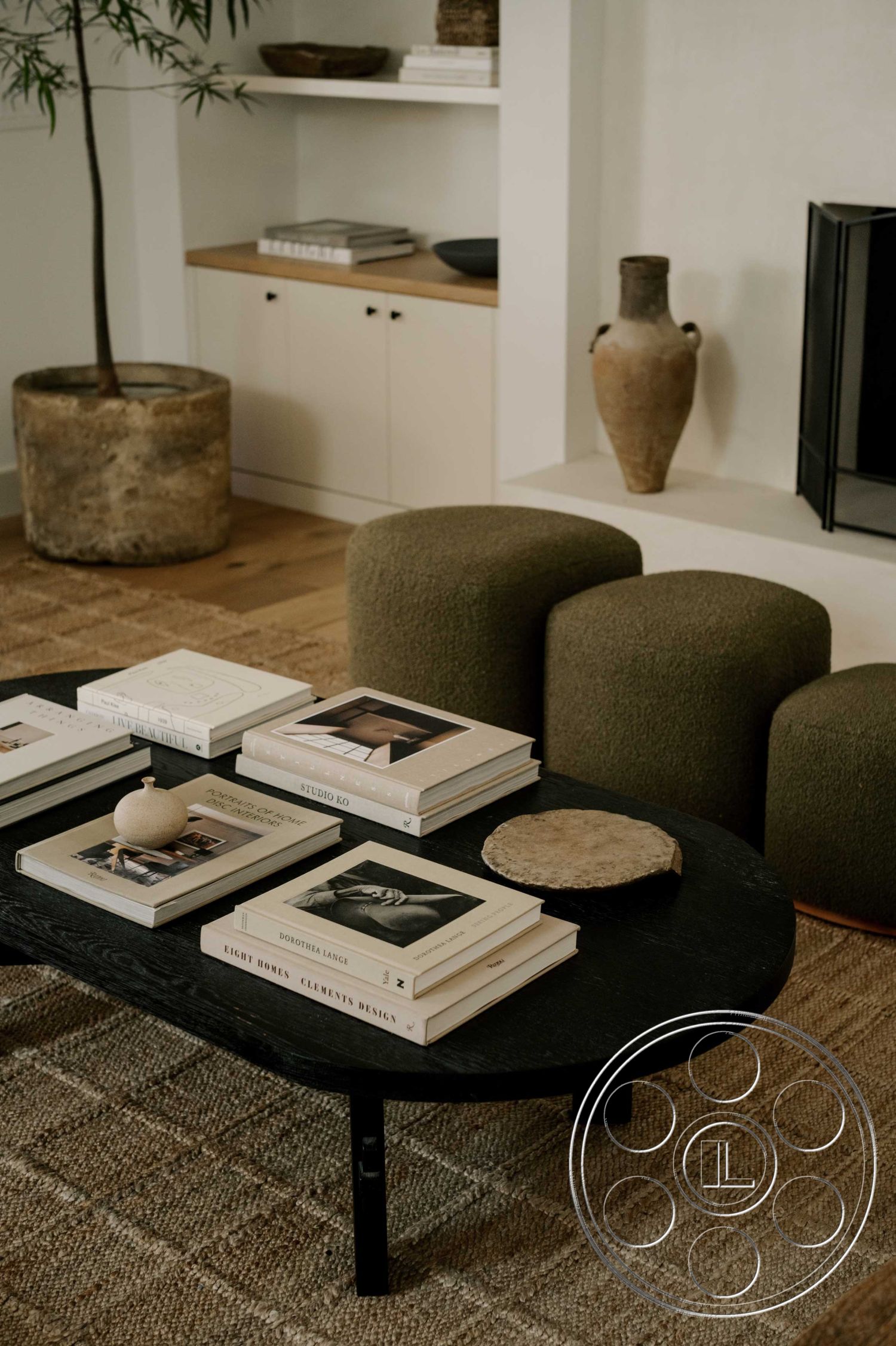 Americana 429 - interior: hardwood flooring, interior: round black wood coffee table, interior: textured area rug, interior: upholstered green ottoman, interior: white painted walls, interior: light wood cabinetry, interior: exposed concrete planter, interior: minimalist decor style, interior: modern accent vases, interior: stacked interior books, interior: clean lines, interior: open shelving, interior: natural light ambiance