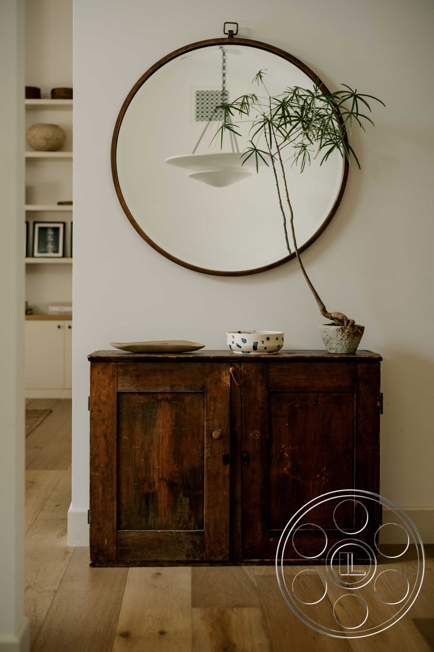 Americana 429 - minimalist decor, earth-toned color palette, interior hardwood flooring, neutral painted walls, vintage wooden cabinet, round wall mirror, green houseplant, ceramic decorative bowl, textured surface decor, light fixtures with minimalist design