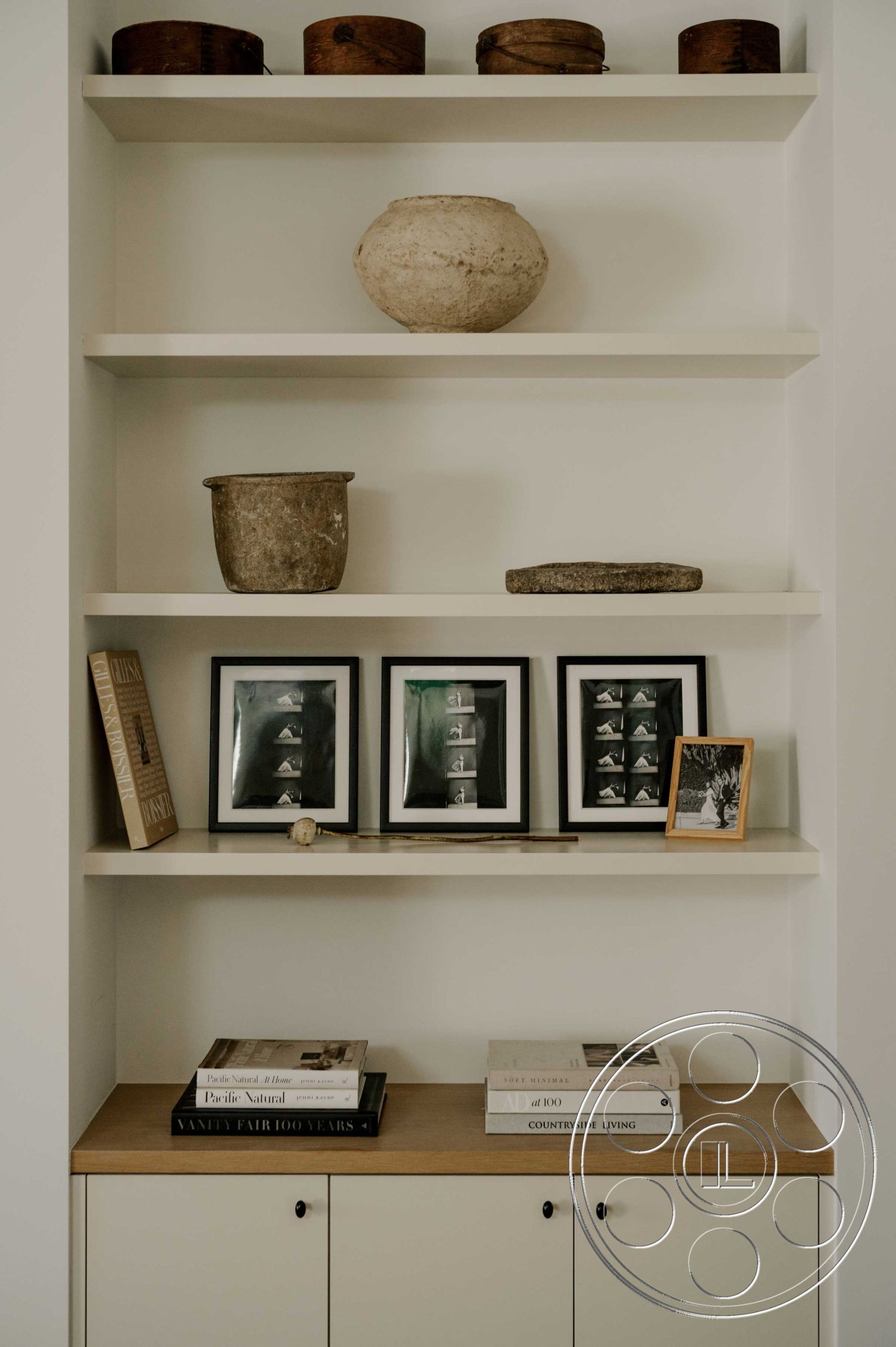 Americana 429 - neutral color palette, natural light illumination, white painted walls, contemporary interior decor, interior shelving unit, natural wood shelf material, ceramic decorative pots, framed black-and-white photographs, textured stone centerpiece, minimalist book display, flat-front cabinet with black hardware, clean lines and modern aesthetic, open shelf design, artistic layering of decorative elements, functional storage space
