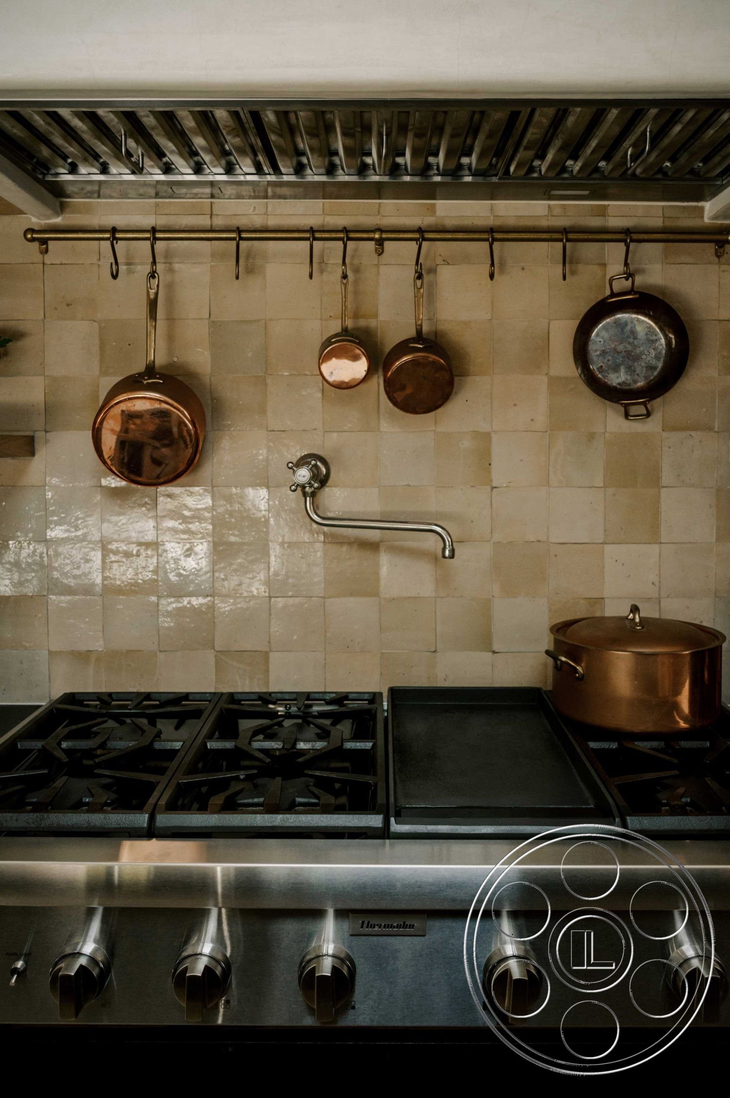 Americana 429 - neutral color palette, dark wood cabinets, modern kitchen design, ceramic tile kitchen floor, stainless steel commercial-grade oven, interior kitchen with concrete tile backsplash, hanging copper cookware, brass pot rack, black cast iron grates on cooktop, custom kitchen shelving, modern faucet with utility sink
