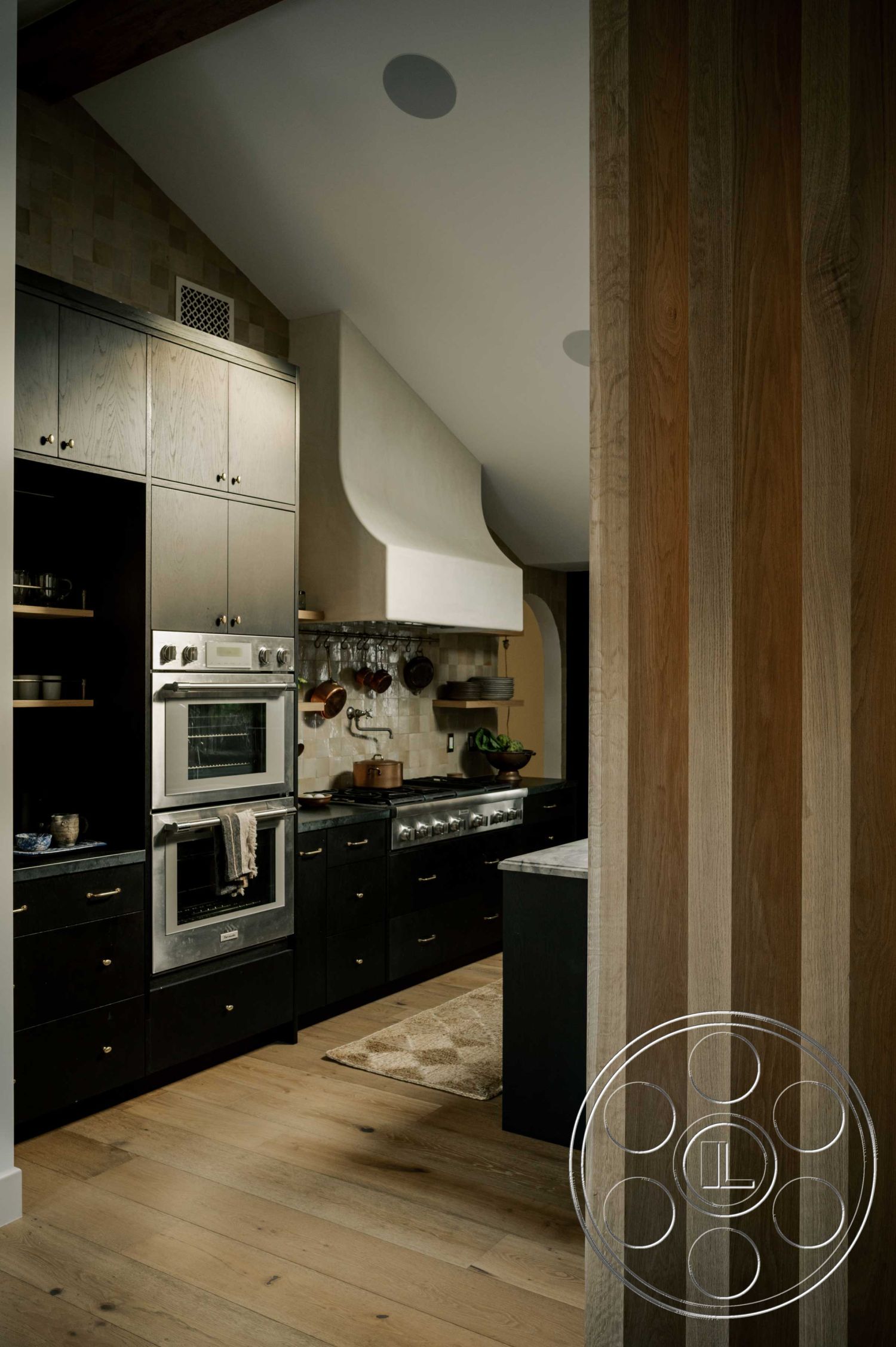 Americana 429 - marble kitchen island countertop, dark wood paneling accent wall, interior kitchen with hardwood flooring, black cabinetry with sleek handles, textured tile kitchen backsplash, built-in stainless steel appliances, high-arched white range hood, open concept kitchen design, natural light from sloped ceiling, modern kitchen with minimalist aesthetics, farmhouse-style kitchen details, subtle area rug on hardwood floor, metal pot rack over kitchen counter, neutral color palette with earthy tones