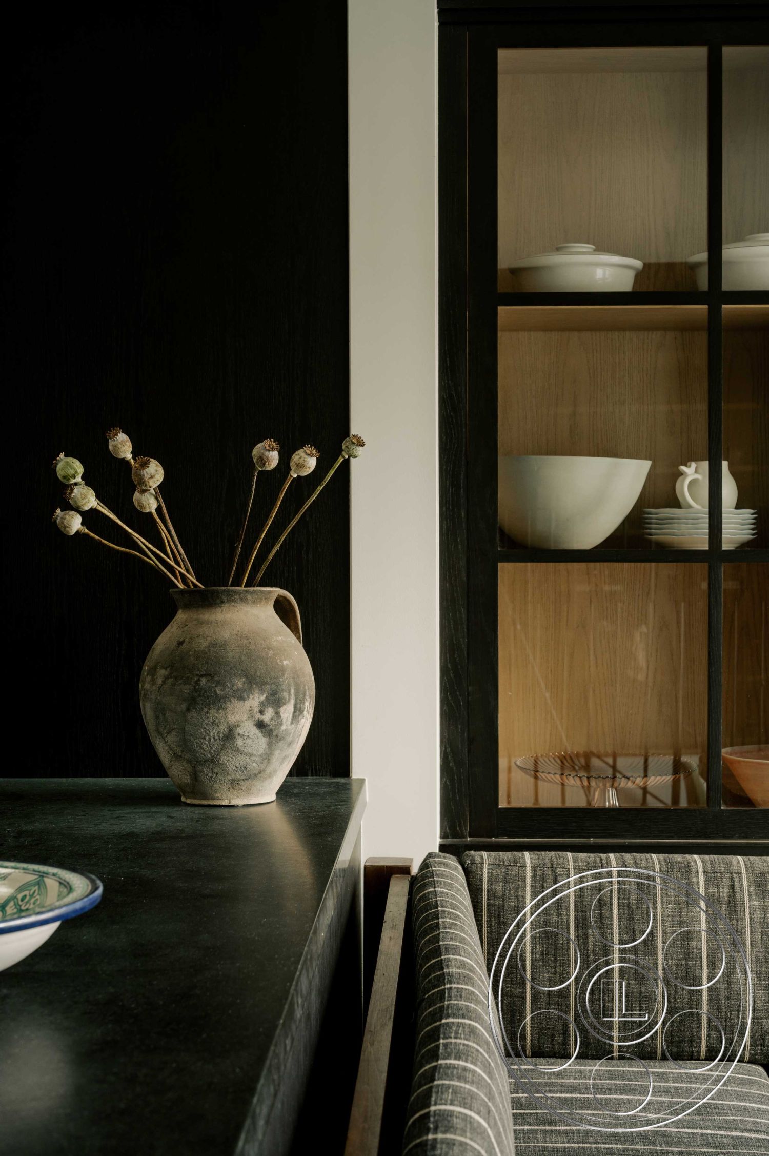 Americana 429 - neutral color palette, light wood cabinetry, contemporary interior design, minimalist kitchen decor, interior kitchen countertop black stone, interior wooden cabinetry with glass doors, striped fabric upholstery on seating, ceramic vase with dried flowers, dark painted accent wall, open shelving design