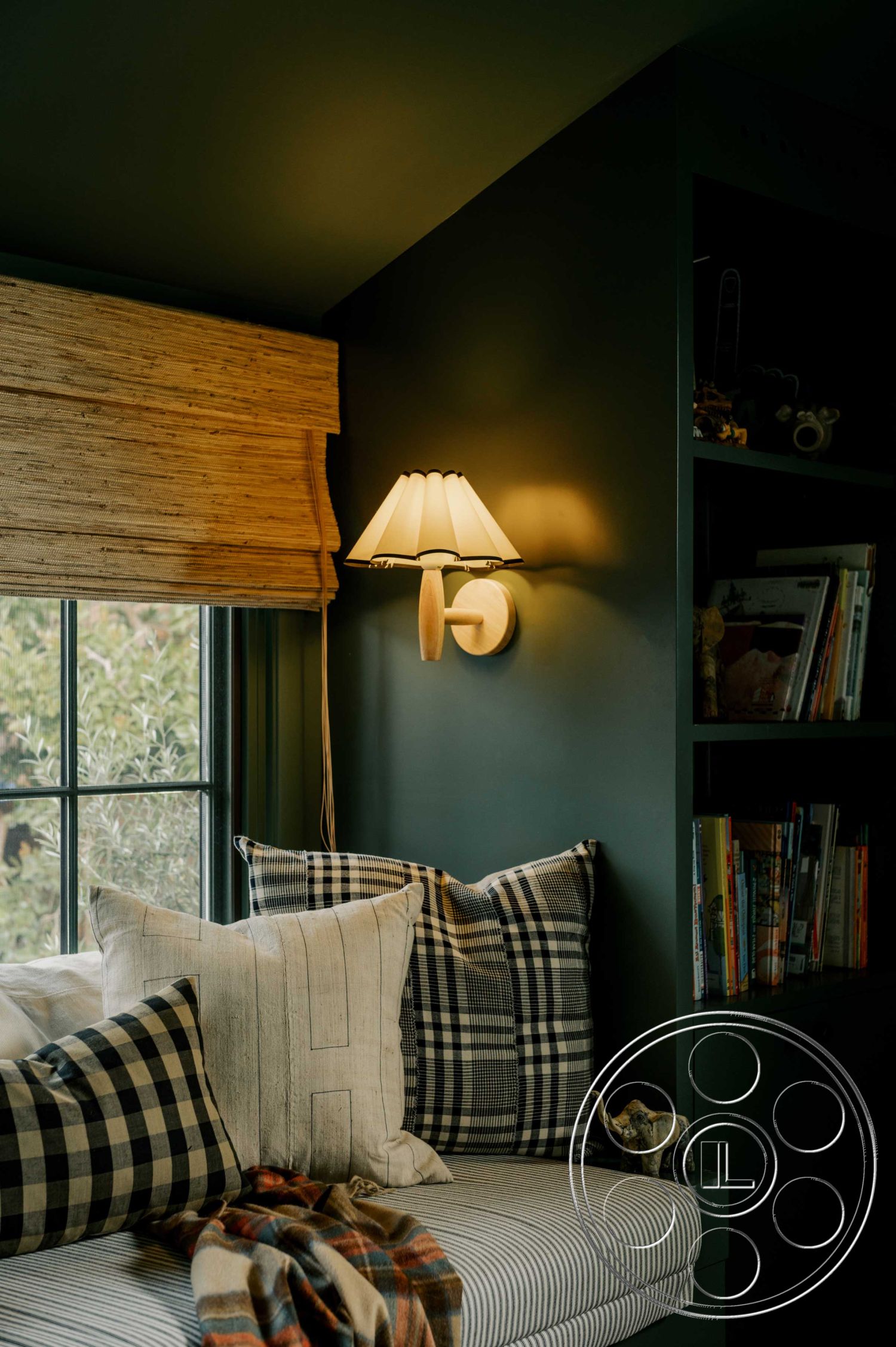 Americana 429 - natural wood accents, built-in bookshelf, modern farmhouse style, plaid throw pillows, striped cushion seat, woven bamboo window shades, dark green painted walls, wall-mounted sconce lighting, textured fabric throws, cozy nook seating area, window with garden view, functional reading corner