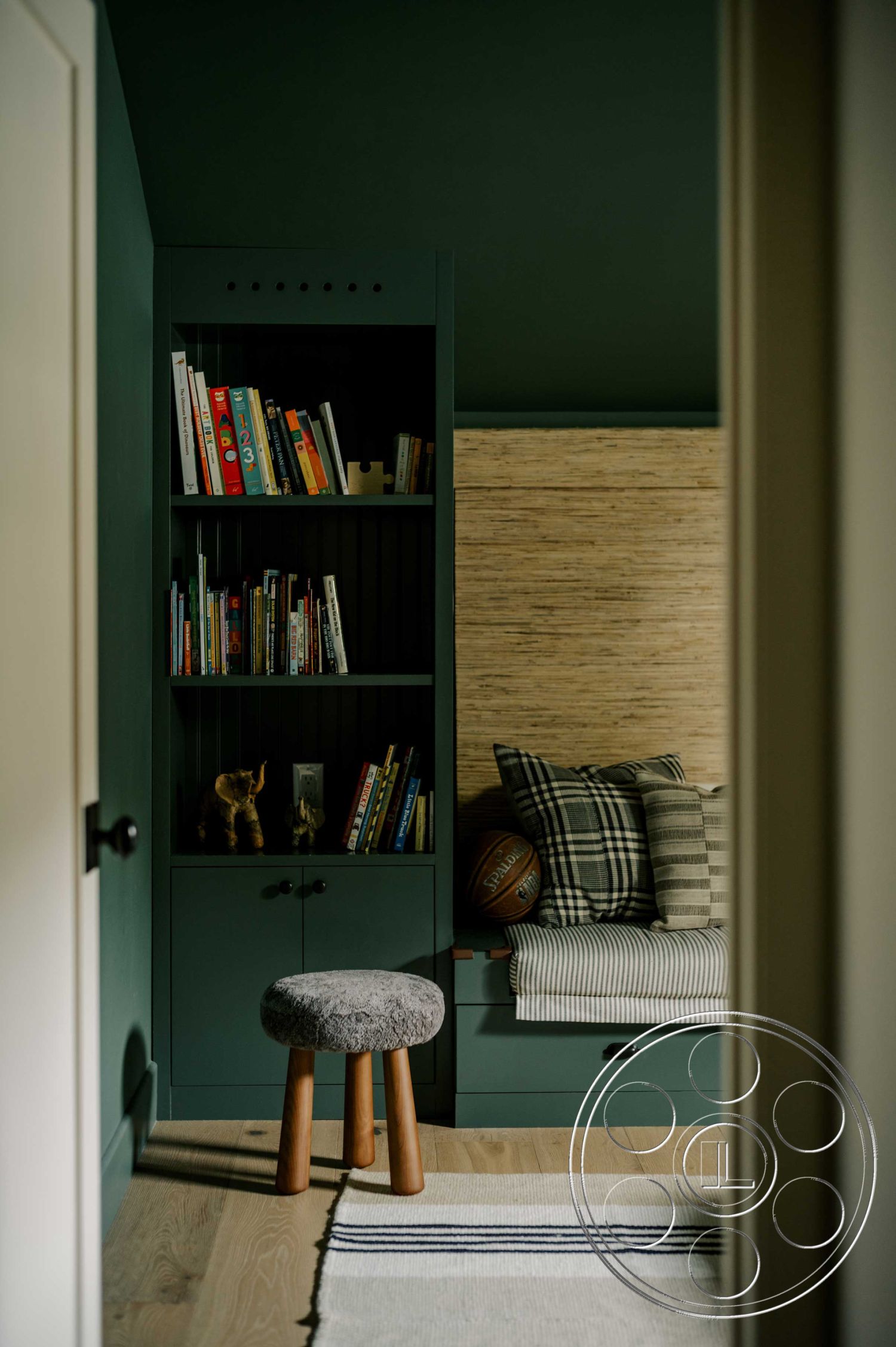 Americana 429 - light hardwood flooring interior, dark green painted walls interior, built-in bookshelves with dark green finish interior, textured natural fiber wall treatment interior, plaid throw pillows on bench seating interior, wooden stool with four legs interior, woven area rug with striped pattern interior, cozy reading nook interior, soft seating area with bench interior, warm, inviting room function interior