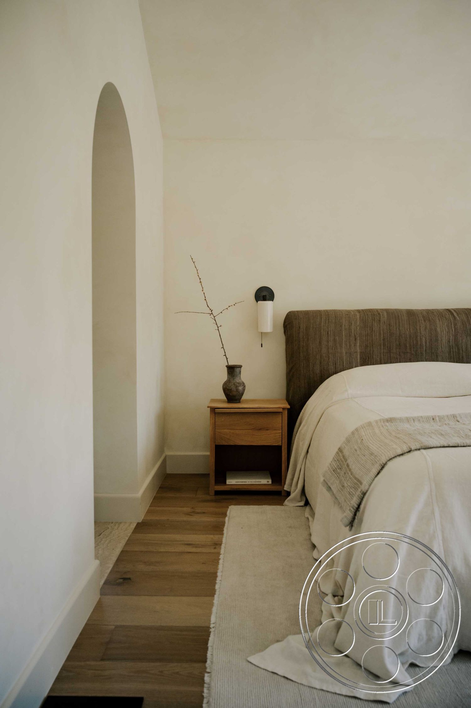 Americana 429 - natural light interior, hardwood bedroom floors interior, neutral color palette interior, exposed wood ceiling beams interior, textured plaster walls interior, arched doorway interior, minimalist wooden nightstand interior, soft linen bedding interior, decorative vase on nightstand interior, woven area rug interior, modern wall sconce lighting interior