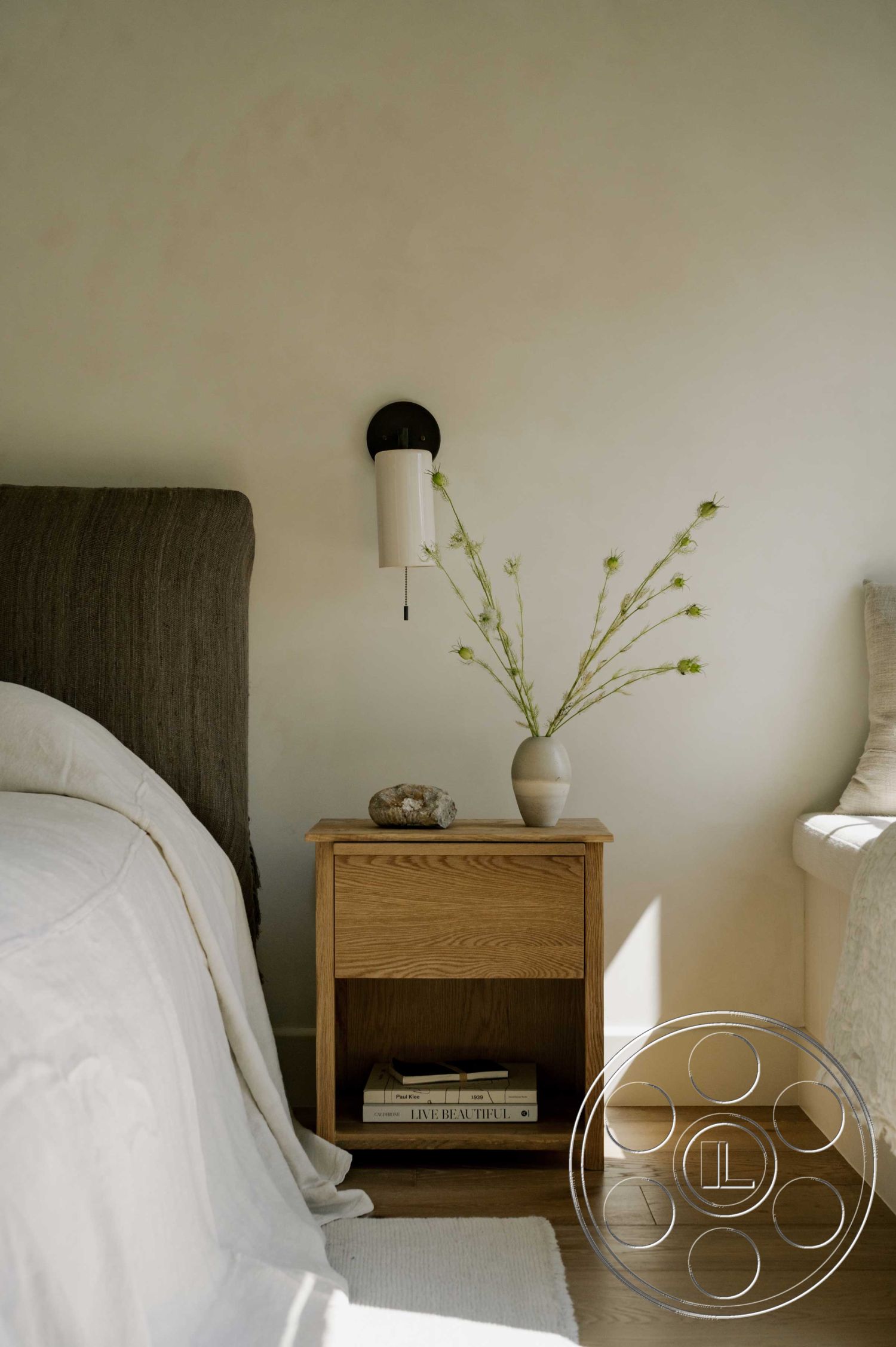 Americana 429 - neutral color palette, cozy reading nook, natural light from window, minimalist design aesthetic, hardwood bedroom floors, wooden nightstand with open shelf, textured plaster bedroom walls, linen bedspread, black wall sconce lighting, ceramic vase on nightstand, decorative stone on nightstand, plant decor with green stems