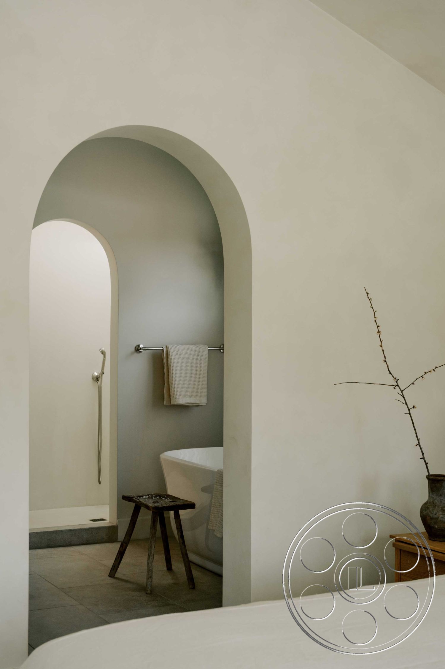 Americana 429 - neutral color palette, spa-like atmosphere, free-standing bathtub, minimalist bathroom design, textured plaster wall treatment, smooth plaster interior walls, arched shower entryway, walk-in shower with minimalist fixtures, natural wood stool, light gray floor tiles, modern interior aesthetic, curved architectural details, contemporary bathroom layout, organic decor elements
