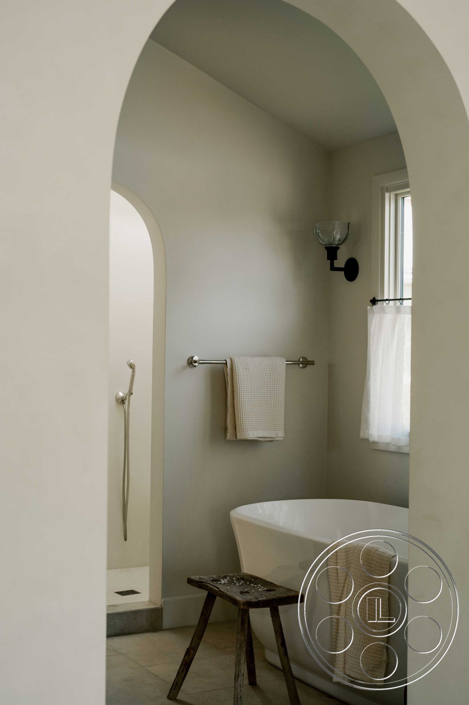 Americana 429 - neutral color palette, large window with sheer curtains, elegant bathroom design, smooth plaster walls, exposed archway interior, freestanding soaking bathtub, natural stone floor tiles, curved shower entrance, wall-mounted towel bar, soft beige bathroom linens, minimalist wooden stool, modern sconces lighting, seamless shower with handheld fixture, cozy bathroom nook