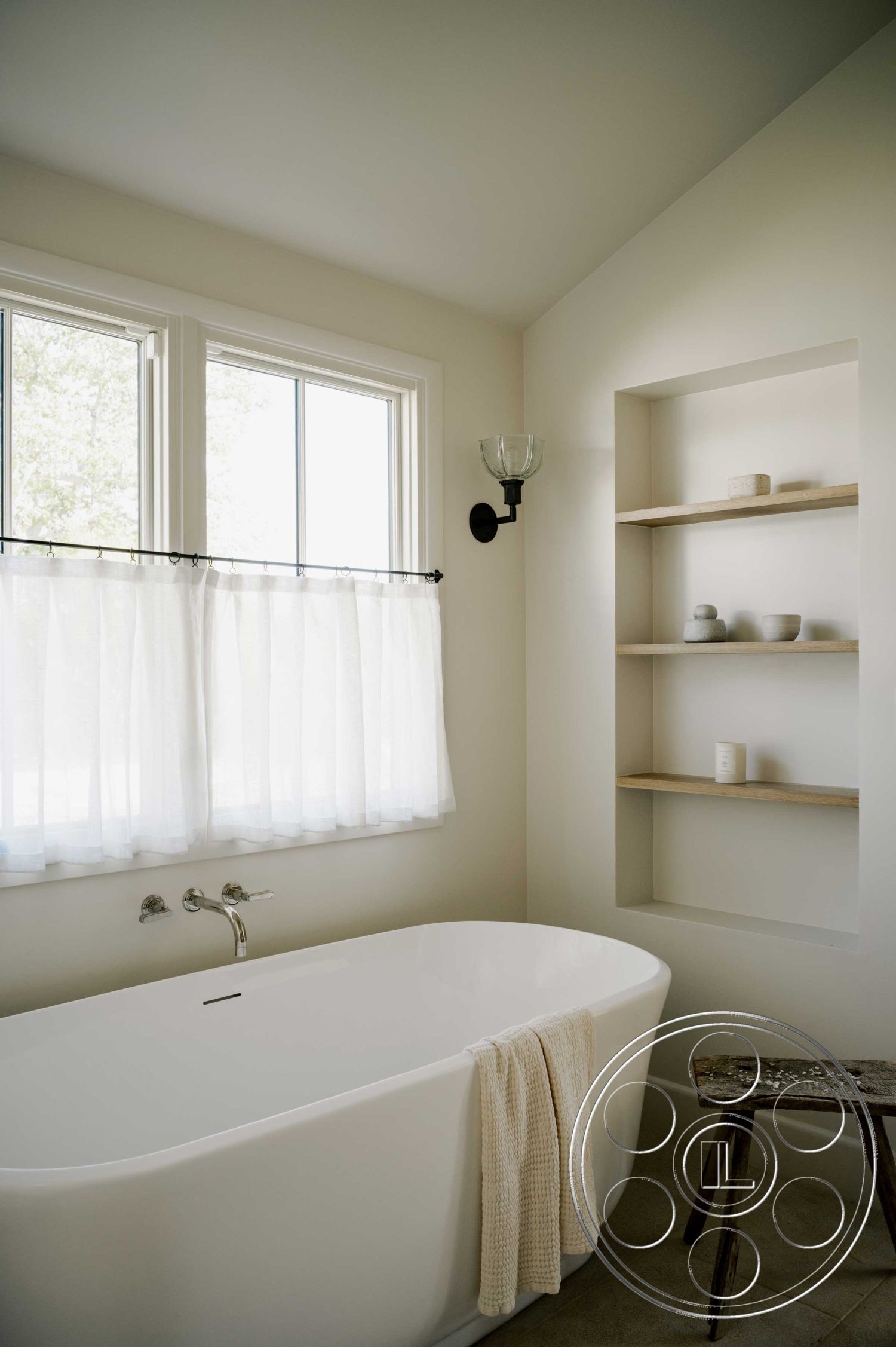 Americana 429 - minimalist bathroom design, neutral tile flooring, decorative wall sconce lighting, textured white plaster walls, freestanding bathtub interior, white ceramic bathtub, stainless steel bathtub faucet, pedestal sink with mirror, glass window with sheer curtains, natural light bathroom setting, wooden floating shelves, soft textile towel, open shelf storage, corner niche for bath accessories