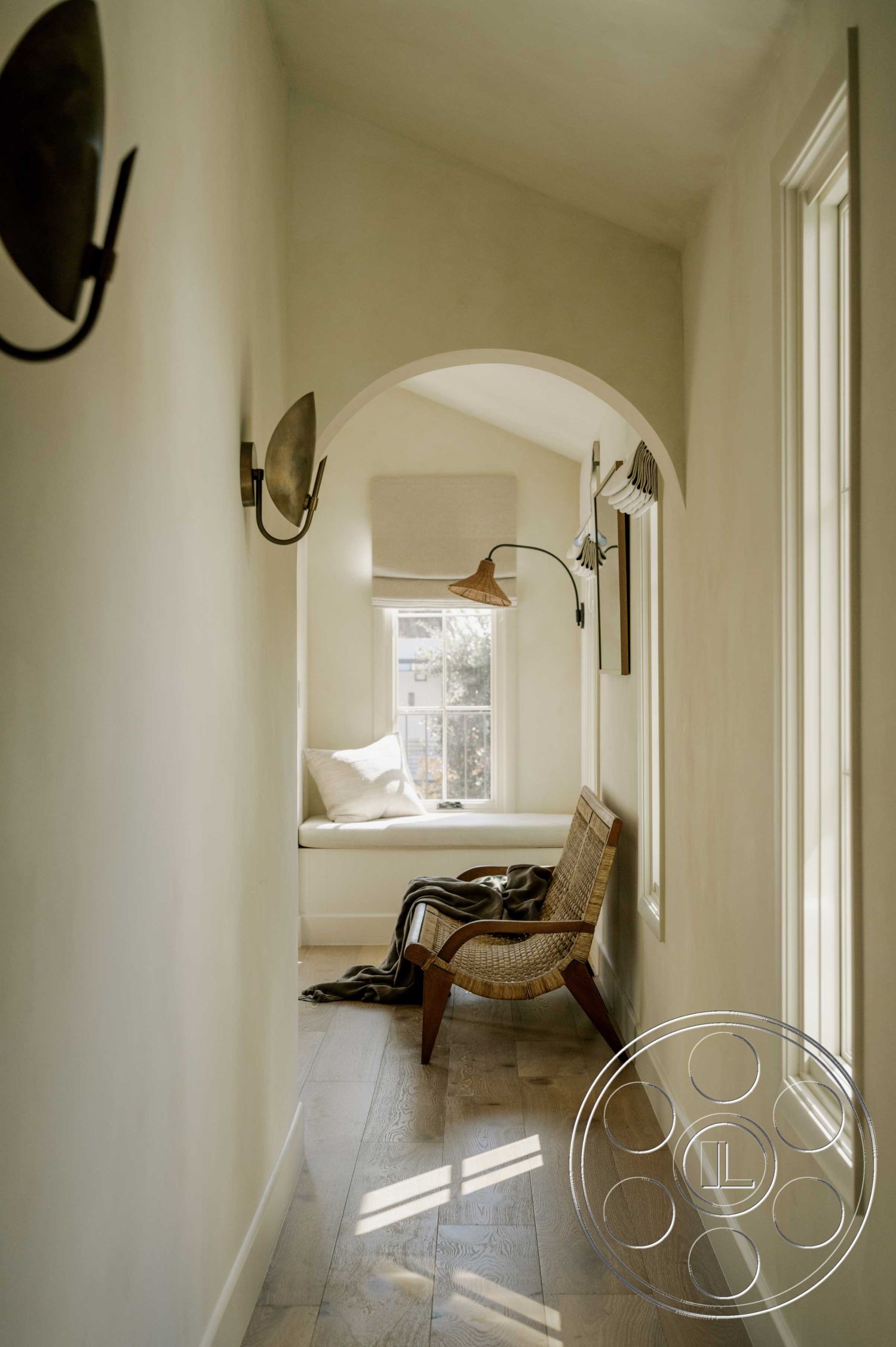 Americana 429 - neutral color palette, natural materials, light hardwood flooring, arched doorways, textured plaster walls, natural light from windows, decorative wall sconces, cozy reading nook, modern minimalist decor, open and airy layout, interior hallway design, built-in bench seat with cushions, woven fabric throw blanket, functionality and aesthetics, traditional window trim